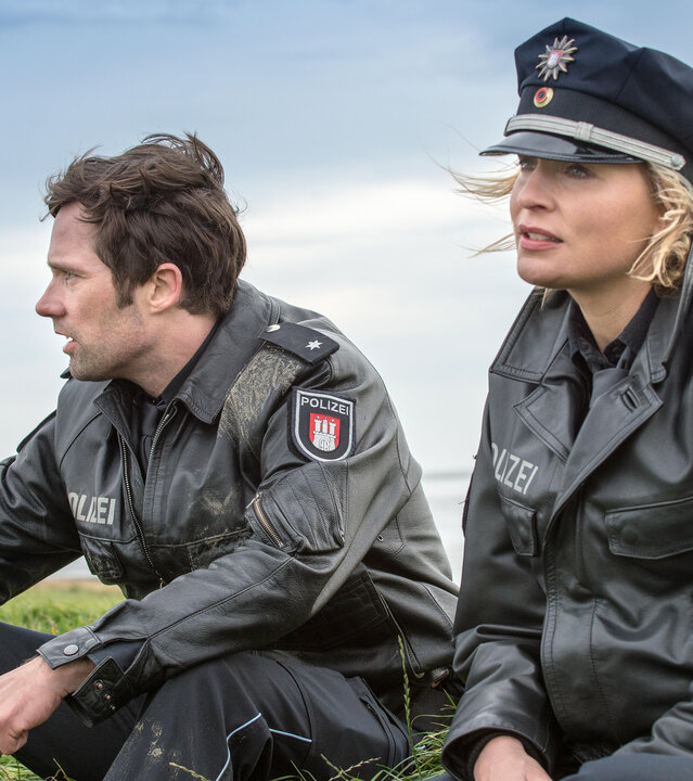 "Notruf Hafenkante - Flucht ins Watt": Die Polizisten Mattes Seeler (Matthias Schloo) und Melanie Hansen (Sanna Englund) sitzen nebeneinander im Gras. Im Hintergrund ist die See zu sehen.