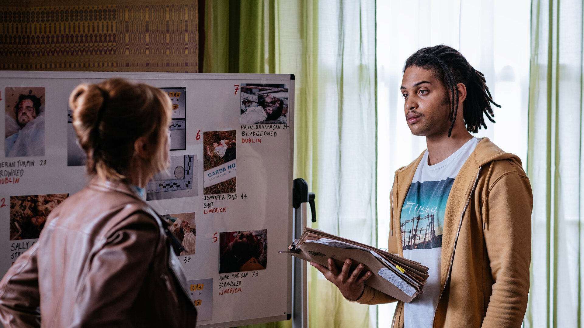 "Harry Wild – Mörderjagd in Dublin - Bei Münzwurf Mord": Harry Wild (Jane Seymour) und Fergus Reid (Rohan Nedd) stehen sich in einem Raum gegenüber. Hinter ihnen ist eine Stellwand mit Fotos und Notizen zu sehen. Fergus hält eine Akte in der Hand.