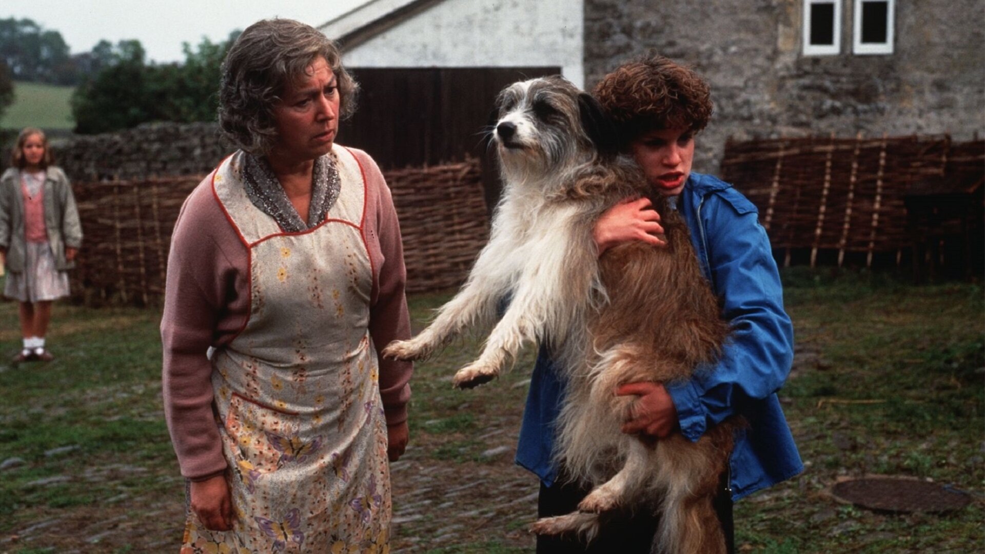 "Fünf Freunde im alten Turm": George (Jemima Rooper) hält den Hund Timmy im Arm, während Mrs. Jones (Ruth Madoc) besorgt auf diesen schaut. Die drei stehen auf einer Wiese. Im Hintergrund steht Anne (Laura Petela) vor einem eingezäunten Haus.