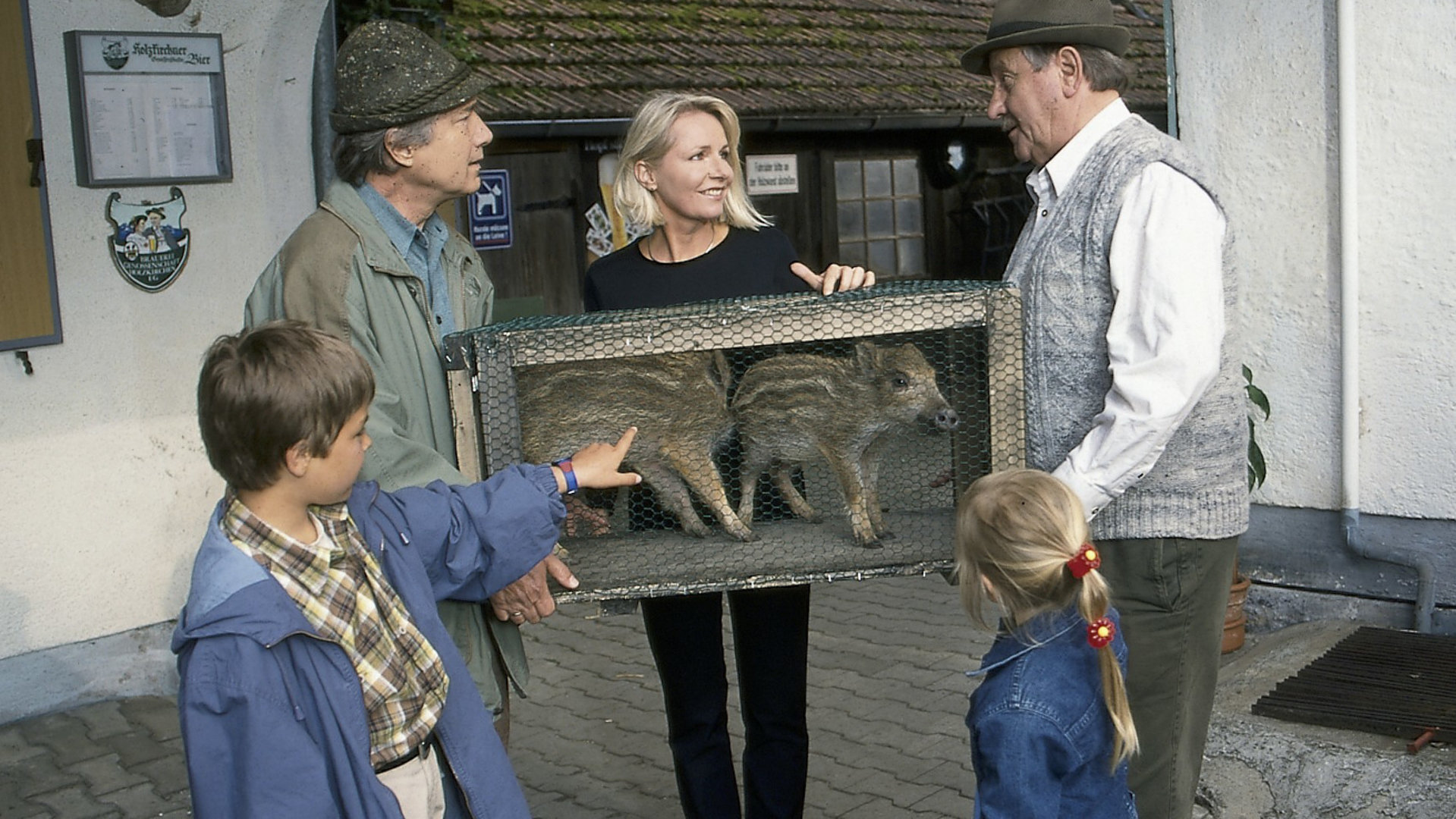 "Forsthaus Falkenau - Eine schöne Schweinerei": Auf einem Bauernhof: Förster Martin Rombach (Christian Wolff), seine Frau Susanna und der kleine Florian (Sascha Hornig) mit seiner Schwester Katharina (Dennis Hornig) stehen mit einer kleine Holzkiste vor dem Bauer Brucks (Gerd Fricks). In der Holzkiste sind zwei Wildschweinfrischlinge.