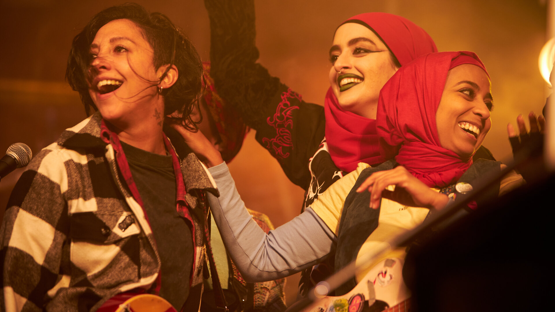 "We are Lady Parts - Sparta": Saira (Sarah Kameela Impey), Ayesha (Juliette Motamed) und Amina (Anjana Vasan) stehen nebeneinander im warmen Licht und lachen gemeinsam. Links neben Saira steht ein Mikrofon.