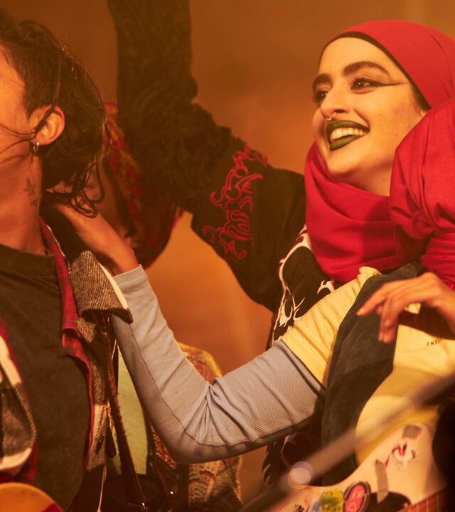 "We are Lady Parts - Sparta": Saira (Sarah Kameela Impey), Ayesha (Juliette Motamed) und Amina (Anjana Vasan) stehen nebeneinander im warmen Licht und lachen gemeinsam. Links neben Saira steht ein Mikrofon.