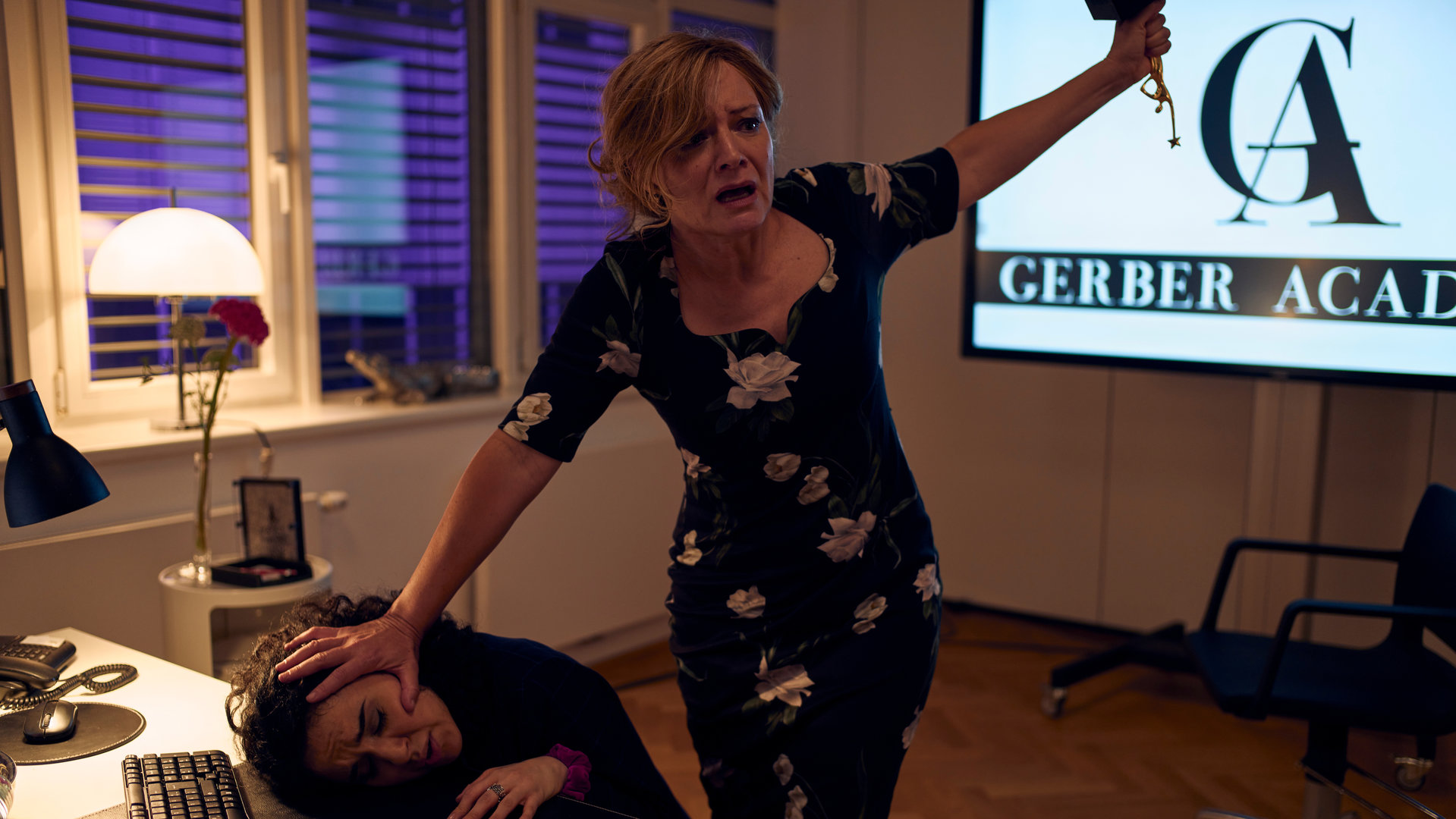 "SOKO Wien - In Schönheit sterben": Ilona Gerber (Marion Mitterhammer) steht hinter Dilaria Yildiz (Mariam Hage) und drückt deren Kopf auf die Schreibtischplatte. Ilona Gerber hat ihren linken Arm ausgestreckt um Schwung zu holen, damit sie Dilaria erschlagen kann.