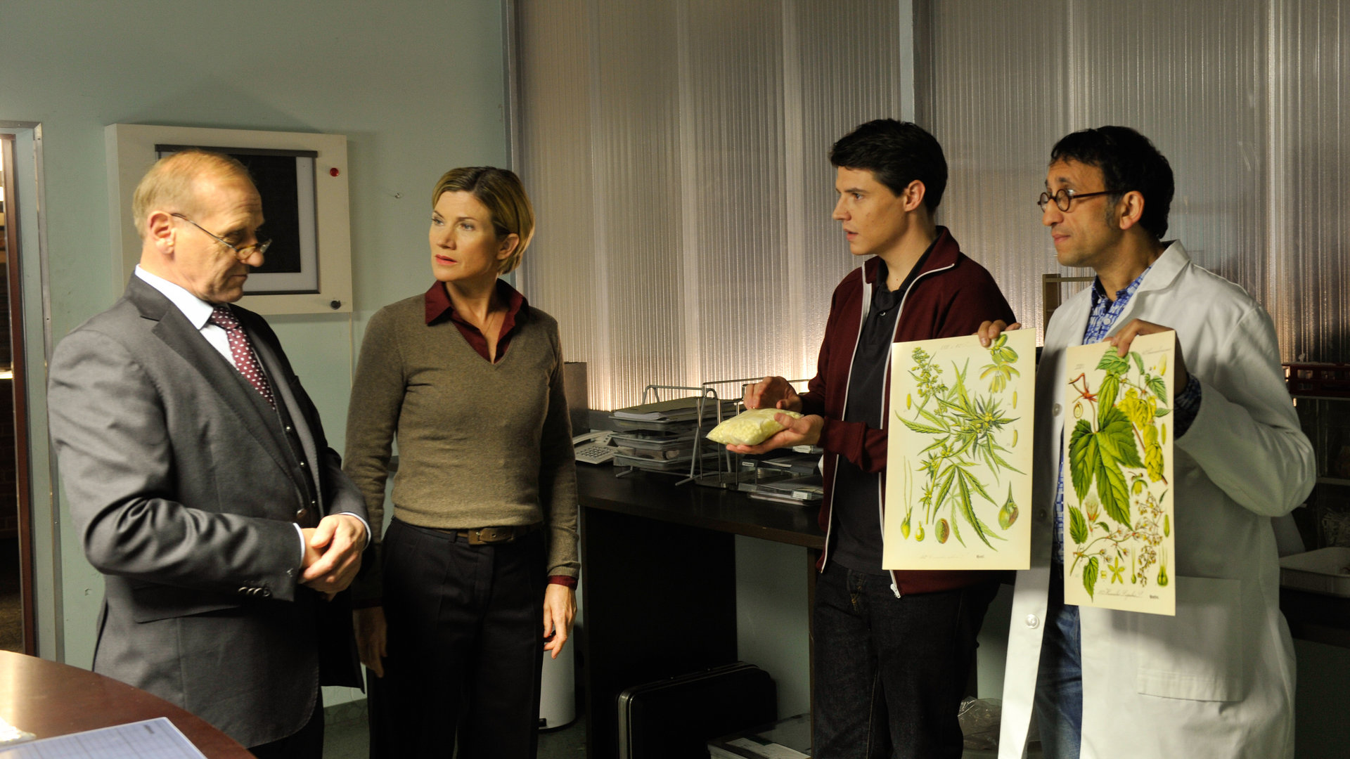 "SOKO Stuttgart - Frauenbier": Kriminaldirektor Michael Kaiser (Karl Kranzkowski) beratschlagt sich mit Martina Seiffert (Astrid M. Fünderich), Rico Sander (Benjamin Strecker) und Jan Arnaud (Mike Zaka Sommerfeldt), der zwei Zeichnungen mit Blättern in die Höhe hält.