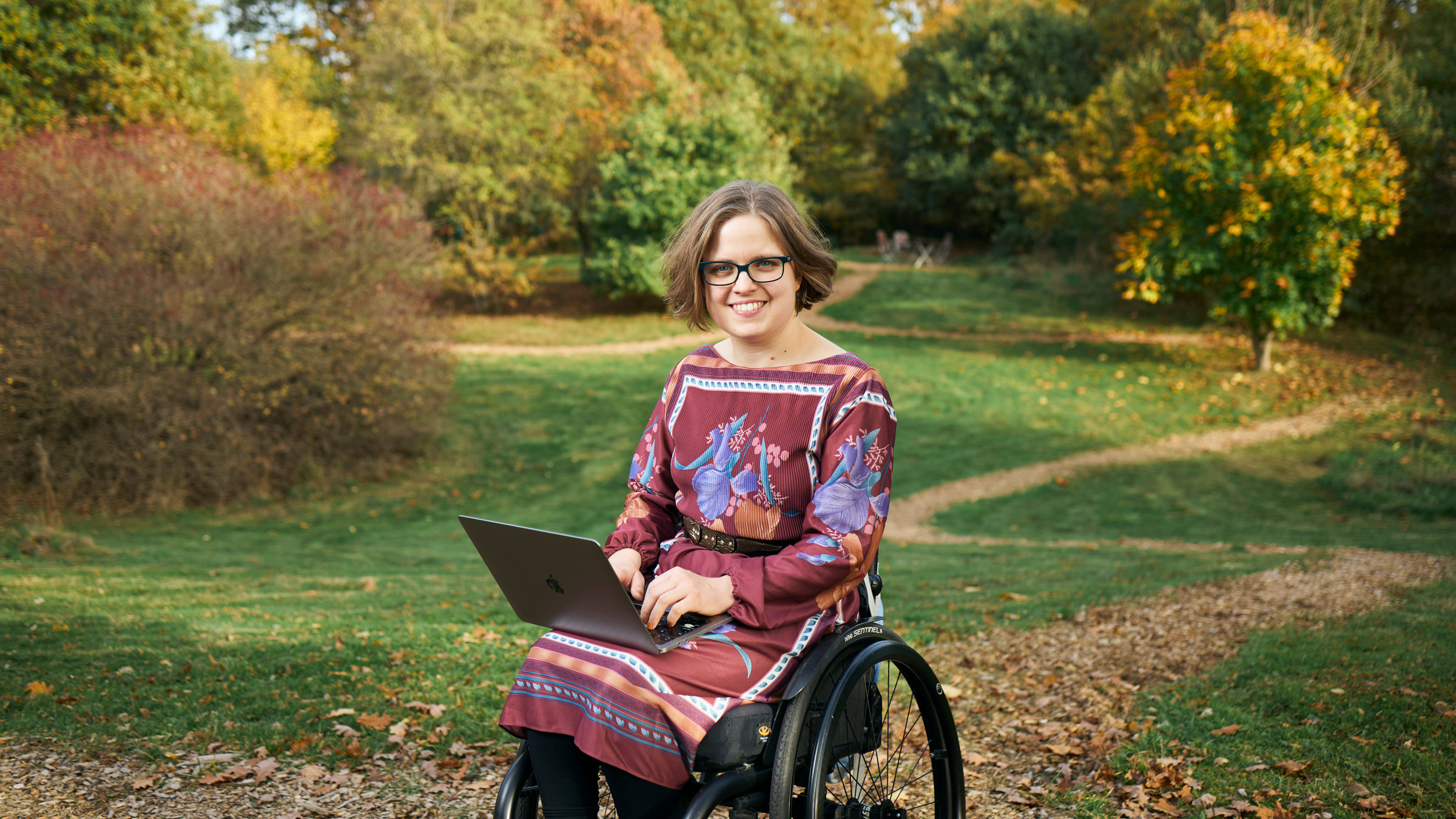 "Einfach Mensch! - Wie wollen wir arbeiten": Cinderella Glücklich sitzt im Rollstuhl im Park. Auf dem Schoß hat sie ein aufgeklapptes Laptop. Beide Hände liegen auf der Tastatur. Sie lächelt in die Kamera.