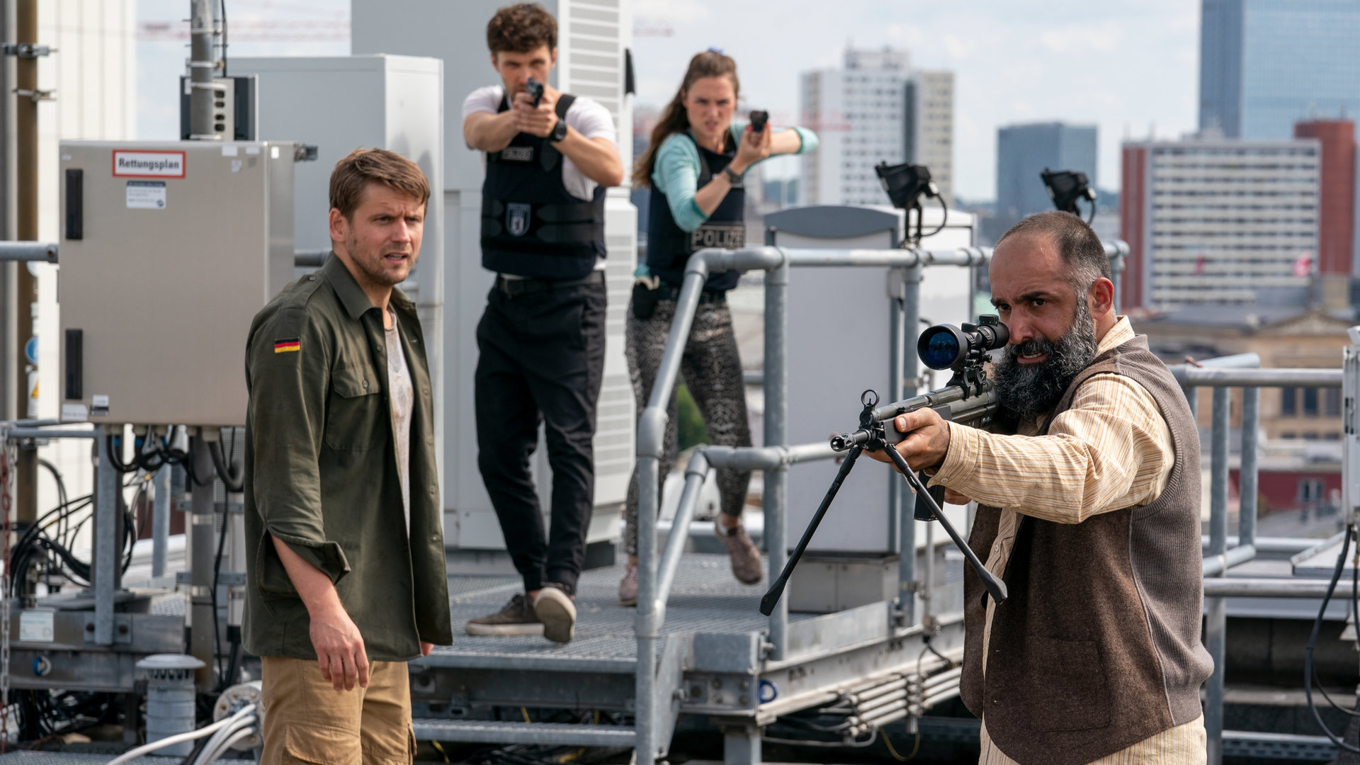 "Letzte Spur Berlin - Tag X - Teil 2": Lucy (Josephin Busch), Alex (Aleksandar Radenković), Mirko Drews (Steve Windolf) und Tahir Adil (David A. Hamade) stehen auf einem Dach. Lucy und Alex richten ihre Waffen auf Tahir Adil, der ebenfalls eine Waffe in der Hand hält und verzweifelt wirkt. Mirko schaut Tahir an.