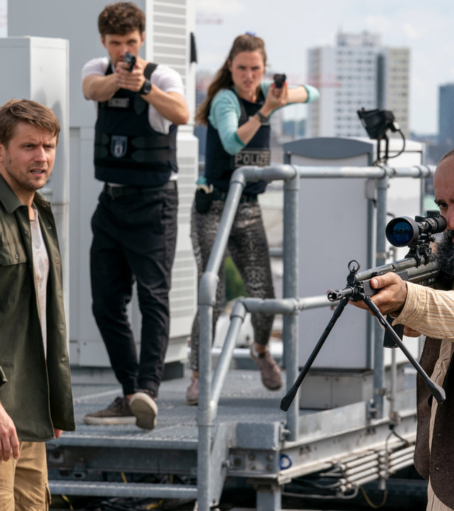 "Letzte Spur Berlin - Tag X - Teil 2": Lucy (Josephin Busch), Alex (Aleksandar Radenković), Mirko Drews (Steve Windolf) und Tahir Adil (David A. Hamade) stehen auf einem Dach. Lucy und Alex richten ihre Waffen auf Tahir Adil, der ebenfalls eine Waffe in der Hand hält und verzweifelt wirkt. Mirko schaut Tahir an.