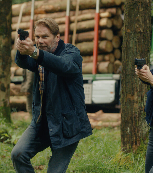 "Erzgebirgskrimi - Mordholz": Im Wald stehen die Ermittler Robert Winkler (Kai Scheve) und Karina Szabo (Lara Mandoki) Seite an Seite mit gezückten Waffen. Ihr Blick richtet sich konzentriert in die gleiche Richtung. Im Hintergrund türmen sich Baumstämme auf einem Lastwagen.
