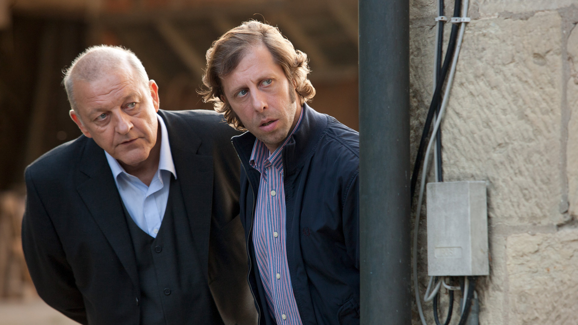 "Wilsberg - Gegen den Strom": Wilsberg (Leonard Lansink) und Ekki (Oliver Korittke) stehen hinter einer Mauer und beobachten etwas.