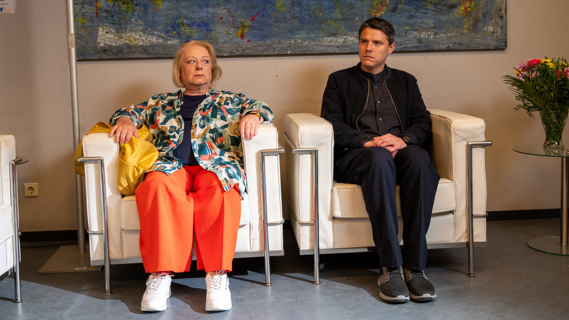 "SOKO Stuttgart - Tod eines Apothekers": Kriminalkommissar Rico Sander (Benjamin Strecker) und seine Mutter Pia (Martina Eitner-Acheampong) sitzen im Wartezimmer eines Krankenhauses nebeneinander auf zwei weißen Ledersesseln. Während Pia ihren Sohn mit ernstem Blick aus dem Augenwinkel beobachtet, schaut dieser angespannt in die Ferne.