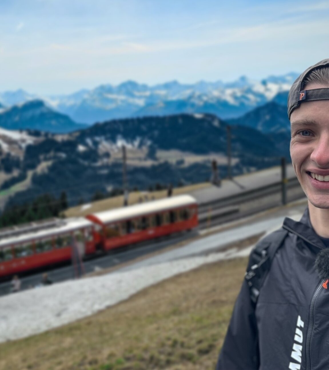 "ZDF.reportage: Reicher Nachbar Schweiz - Leben und Arbeiten in der Grenzregion":  Niklas steht in den Schweizerbergen, hinter ihm fährt eine Bergbahn. Dahinter liegt ein Alpenpanorama.