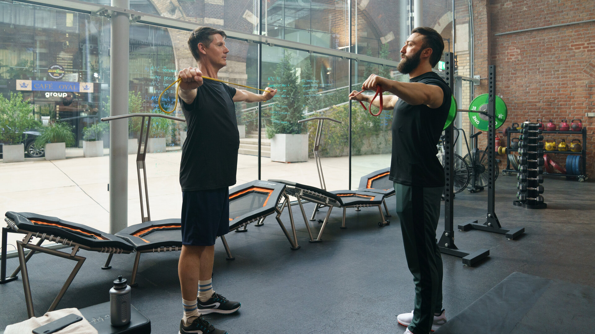 "SOKO Wien - Sisyphos": Thomas Albrechts (Thomas Clemens) steht seinem Fitnesstrainer Masud Ansari (Aslan Aslan) mitten im Fitnessstudio gegenüber. Beide haben die Arme seitlich ausgestreckt und halten ein Gummiband gespannt.  Drumherum stehen verschiedene Fitnessgeräte. Hinter Thomas Albrechts kann man durch eine Glaswand auf eine Einkaufspassage blicken.