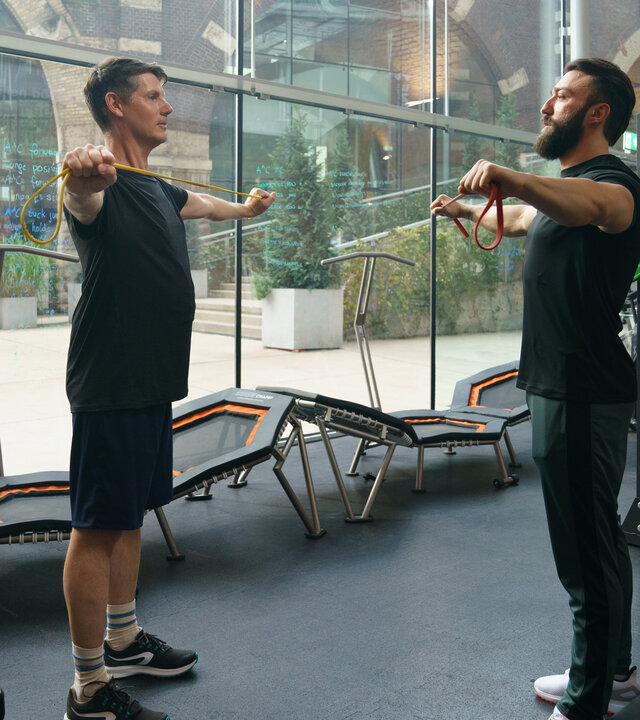 "SOKO Wien - Sisyphos": Thomas Albrechts (Thomas Clemens) steht seinem Fitnesstrainer Masud Ansari (Aslan Aslan) mitten im Fitnessstudio gegenüber. Beide haben die Arme seitlich ausgestreckt und halten ein Gummiband gespannt.  Drumherum stehen verschiedene Fitnessgeräte. Hinter Thomas Albrechts kann man durch eine Glaswand auf eine Einkaufspassage blicken.