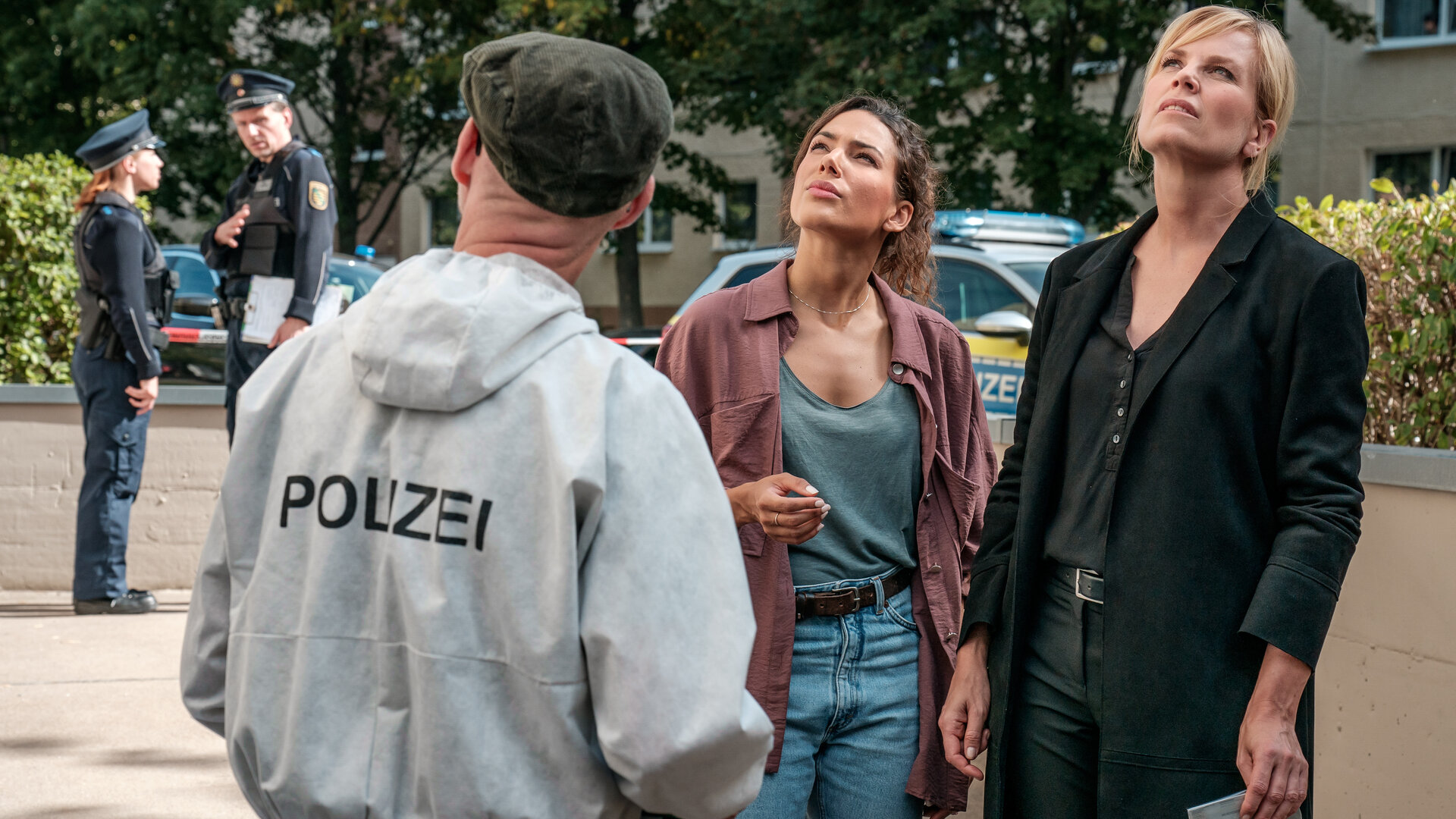 "SOKO Leipzig - Die Ansage": Kim Nowak (Amy Mußul) und Ina Zimmermann (Melanie Marschke) stehen nebeneinander auf dem Gehweg und schauen fragend nach oben auf das Dach. Dabei hat Ina einen Personalausweis in einem Plastiktütchen in der Hand. Vor ihnen steht Holger (Holger Koch) von der KTU, im Hintergrund zwei Personen von der Polizei, die sich unterhalten
