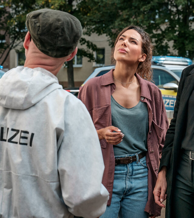 "SOKO Leipzig - Die Ansage": Kim Nowak (Amy Mußul) und Ina Zimmermann (Melanie Marschke) stehen nebeneinander auf dem Gehweg und schauen fragend nach oben auf das Dach. Dabei hat Ina einen Personalausweis in einem Plastiktütchen in der Hand. Vor ihnen steht Holger (Holger Koch) von der KTU, im Hintergrund zwei Personen von der Polizei, die sich unterhalten