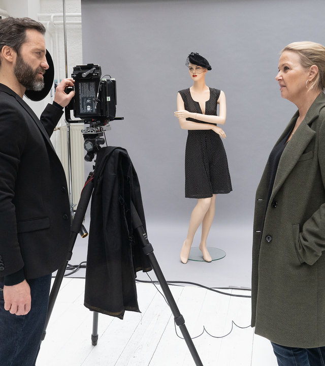 "Die Chefin - True Crime": In einem weißen Fotostudio befragt Vera Lanz (Katharina Böhm) den schwarz gekleideten Fotografen Ulf Brandt (Thomas Wodianka). Er hat eine Hand an seiner Kamera, die auf einem Stativ steht und auf eine ausgerollte graue Leinwand gerichtet ist. Dort steht eine Schaufensterpuppe mit verschränkten Armen, gekleidet in ein kurzes schwarzes Kleid und eine schwarze Baskenmütze.
