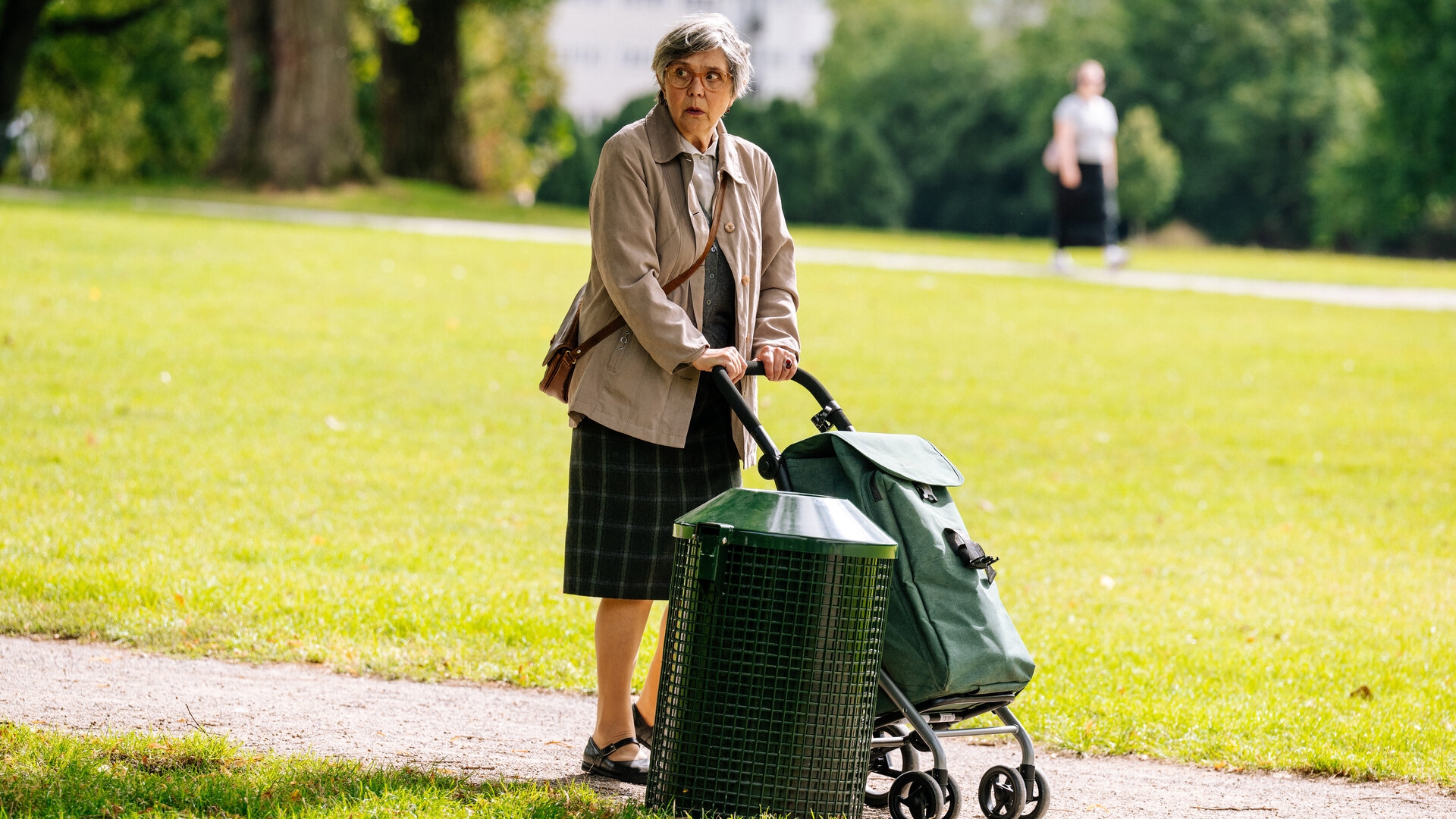 "SOKO Köln - Lilly und Frau Thalheim": Johanna Thalheim (Doris Plenert) steht auf einem Weg in einem Park neben einem grünen Mülleimer. Sie schiebt einen grünen Einkaufstrolley vor sich her und blickt aufgeregt nach links, an der Kamera vorbei. Im Hintergrund sieht man verschwommen eine weitere Person (Komparse) durch den Park laufen.