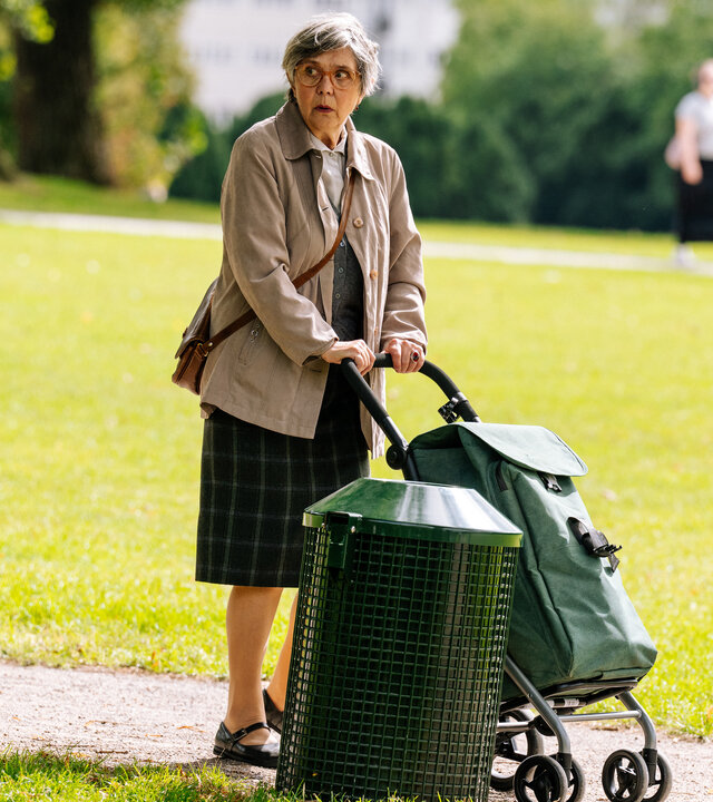 "SOKO Köln - Lilly und Frau Thalheim": Johanna Thalheim (Doris Plenert) steht auf einem Weg in einem Park neben einem grünen Mülleimer. Sie schiebt einen grünen Einkaufstrolley vor sich her und blickt aufgeregt nach links, an der Kamera vorbei. Im Hintergrund sieht man verschwommen eine weitere Person (Komparse) durch den Park laufen.