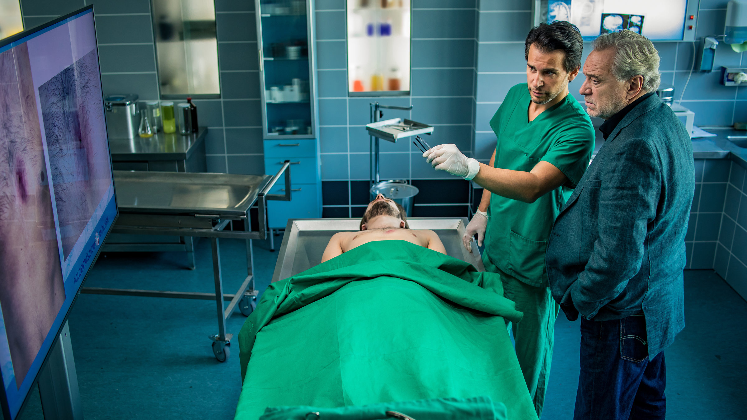 "SOKO München - Fischerstechen": Arthur Bauer (Gerd Silberbauer) steht neben Dr. Weissenböck (Florian Odendahl) in der Pathologie. Vor ihnen auf dem Sektionstisch liegt die Leiche eines Mannes. Der Oberkörper des Toten ist nackt. Von den Füßen bis zu seinem  Bauch wurde ihm eine grüne Decke über den Körper gelegt. Dr. Weissenböck trägt eine grüne Arztkleidung und weiße Gummihandschuhe. In seiner linken Hand hält er eine Pinzette mit der er auf einen großen Bildschirm deutet, der ihnen gegenüber steht. Sie betrachten nachdenklich das Bild auf dem Monitor. Auf diesem sieht man den nackten Oberkörper des Opfers mit einer ovalen Wunde im Brustbereich.