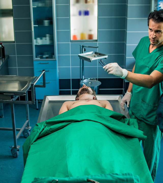 "SOKO München - Fischerstechen": Arthur Bauer (Gerd Silberbauer) steht neben Dr. Weissenböck (Florian Odendahl) in der Pathologie. Vor ihnen auf dem Sektionstisch liegt die Leiche eines Mannes. Der Oberkörper des Toten ist nackt. Von den Füßen bis zu seinem  Bauch wurde ihm eine grüne Decke über den Körper gelegt. Dr. Weissenböck trägt eine grüne Arztkleidung und weiße Gummihandschuhe. In seiner linken Hand hält er eine Pinzette mit der er auf einen großen Bildschirm deutet, der ihnen gegenüber steht. Sie betrachten nachdenklich das Bild auf dem Monitor. Auf diesem sieht man den nackten Oberkörper des Opfers mit einer ovalen Wunde im Brustbereich.