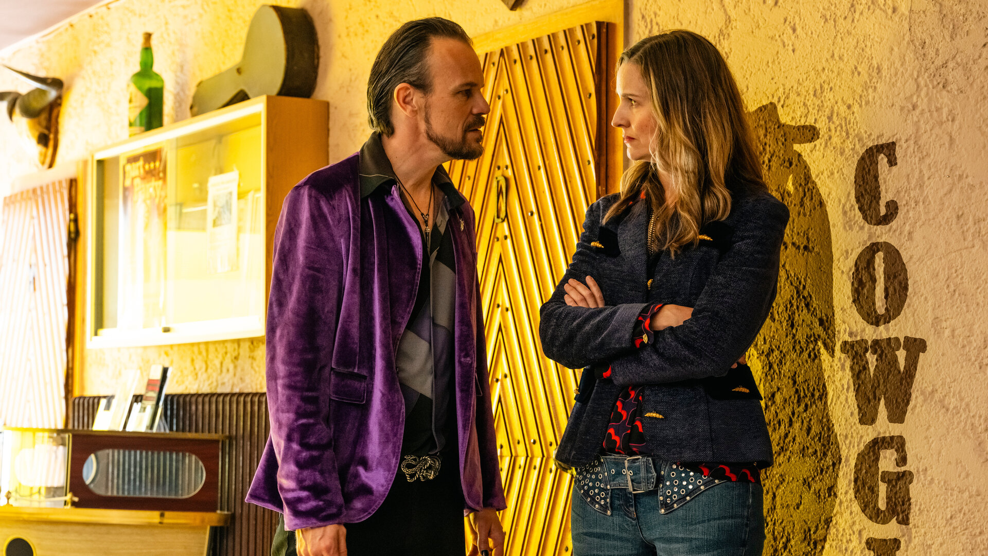 "SOKO Köln - Dart King": In einem Innenraum: Sascha Schuster (Robert Besta) steht vor Heike König (Steffie Neubauer) und sieht sie ernst an. Diese hat die Arme vor der Brust verschränkt und sieht Sascha Schuster wütend an. Das Bild ist in gelbes Licht getaucht.