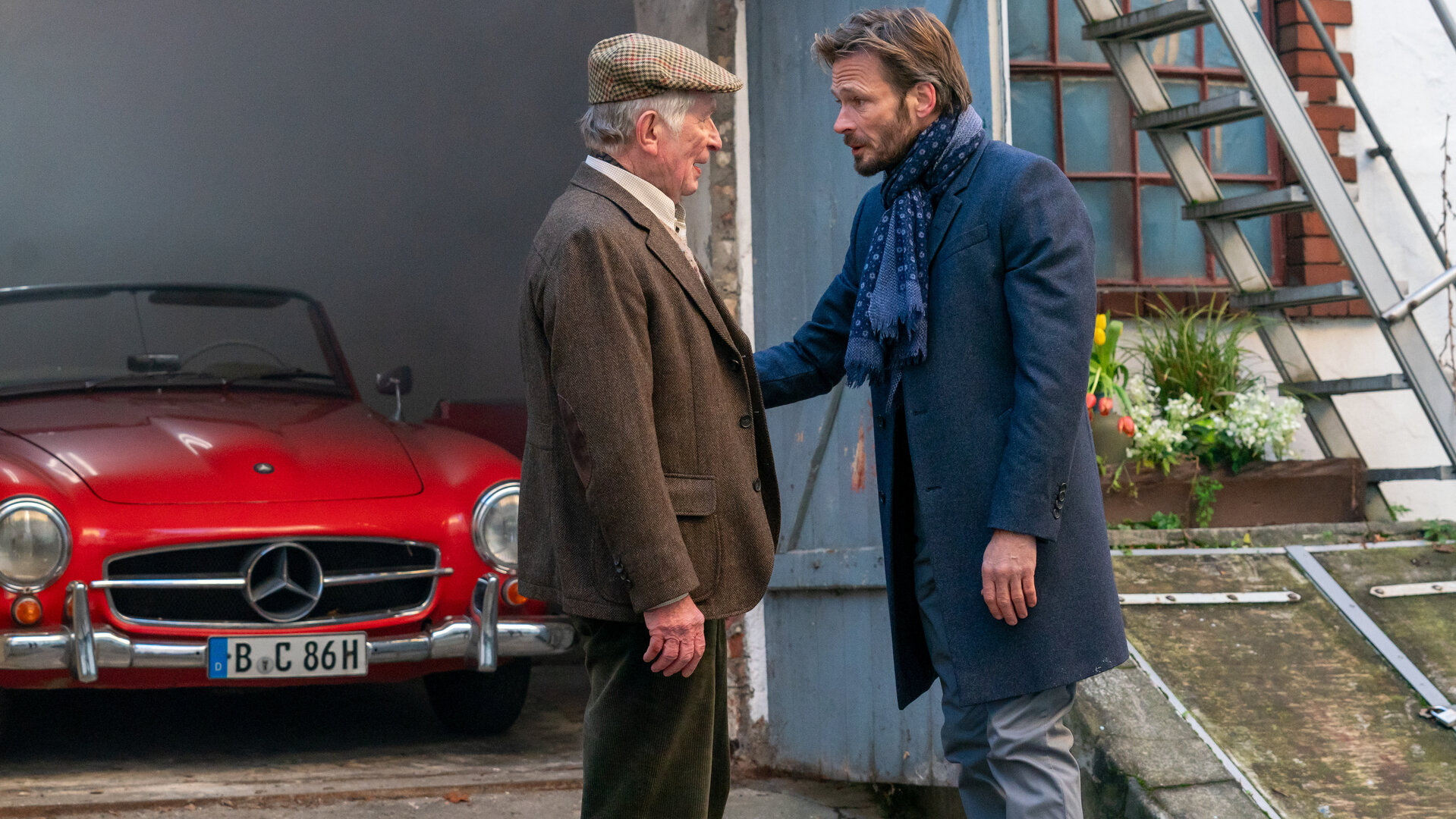 "Letzte Spur Berlin - Ausgeliefert": Otto Haverkamp (Hermann Beyer) steht barfuß vor seinem Sohn Ulf Haverkamp (Andreas Pietschmann) der liebevoll seinen Arm hält. Hinten ist ein schicker, roter Oldtimer in einer offenen Garage geparkt.