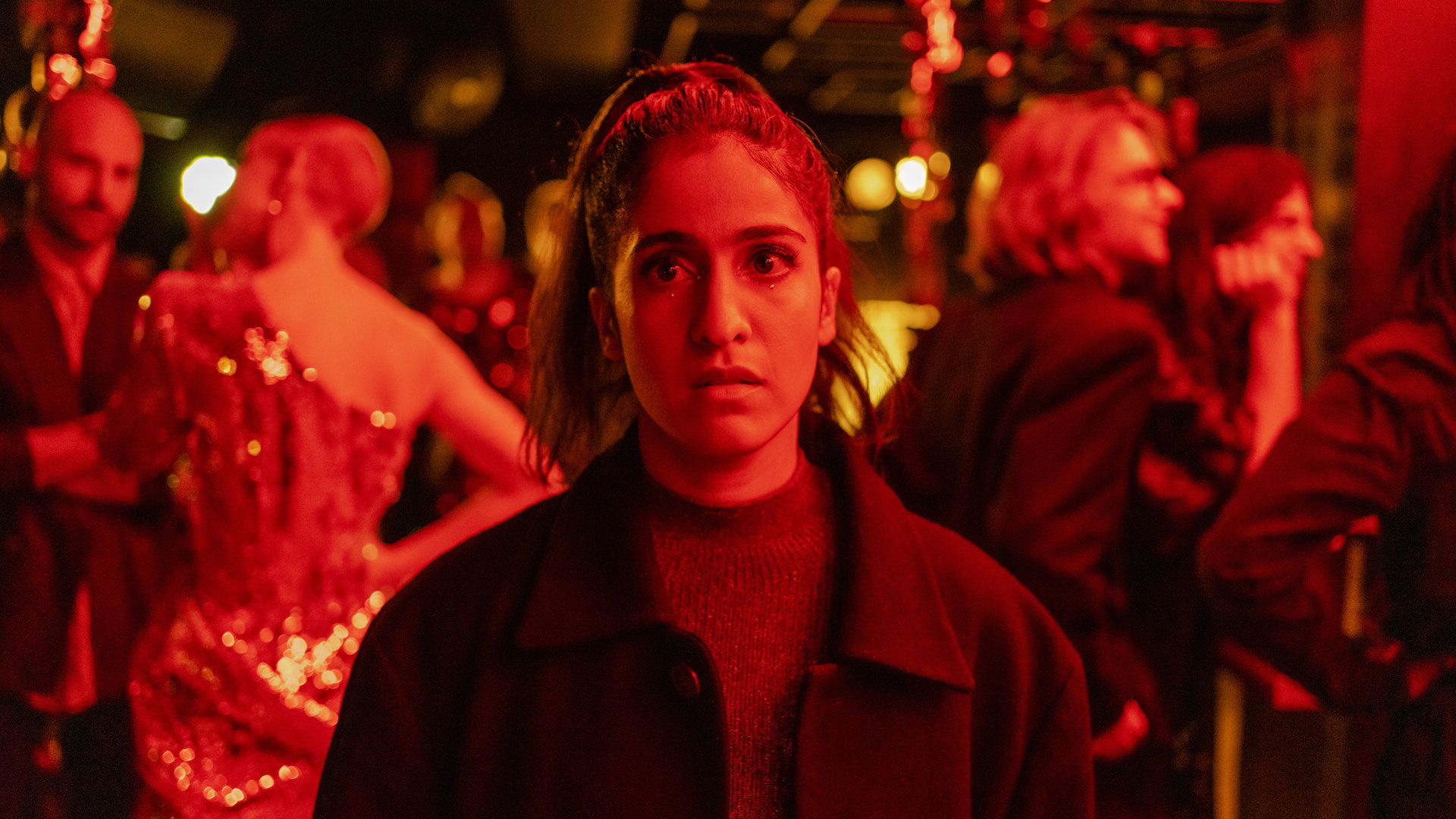 "Loving Her - Loving Truth": Großaufnahme von Hanna (Banafshe Hourmazdi). Sie steht in einem Club. Hinter ihr stehen Menschen (Komparsen), die sich miteinander unterhalten. Sie schaut nachdenklich ins Leere.