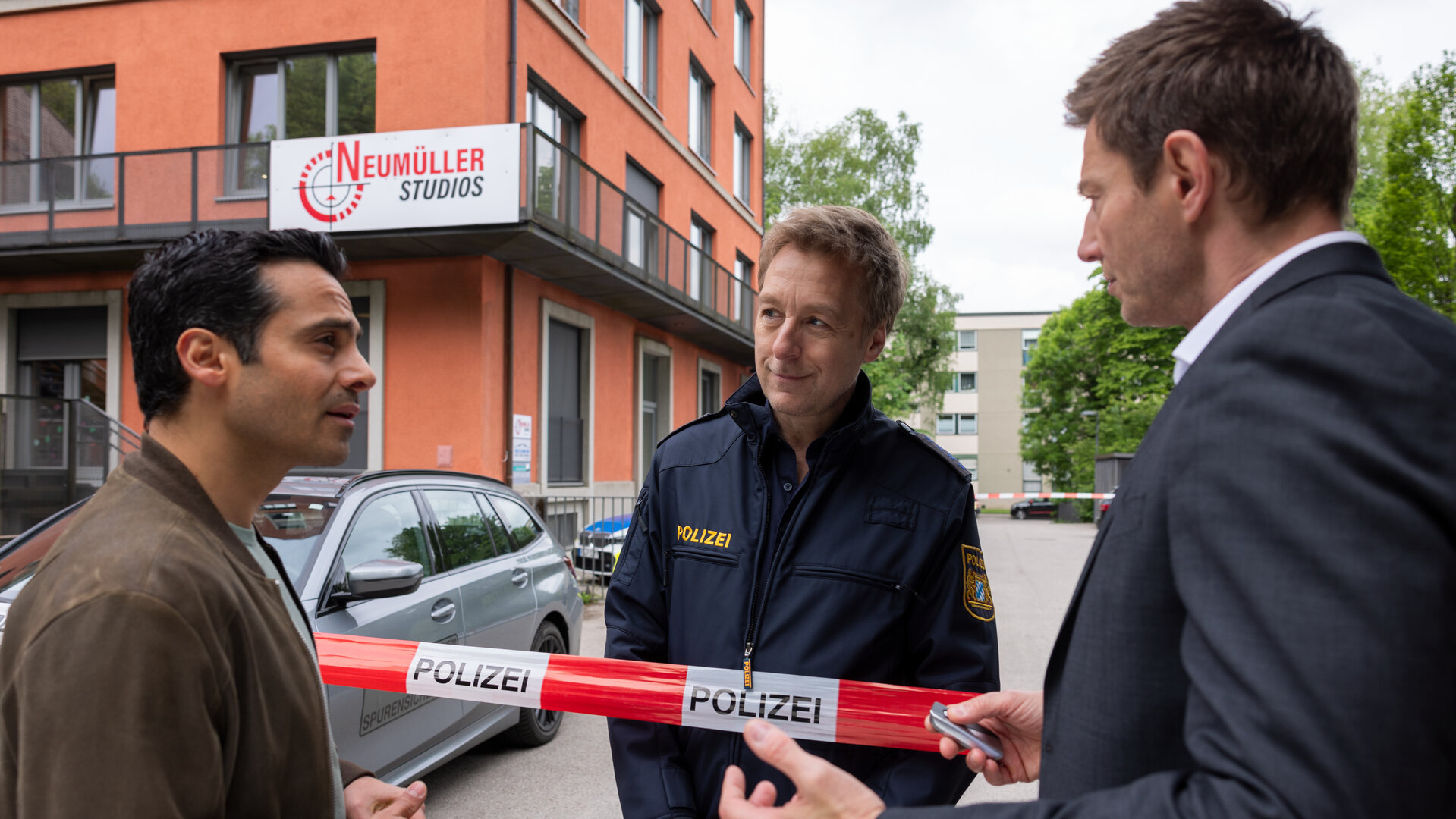 "Die Rosenheim-Cops - Die Autogrammjagd": Kommissar Kaya (Baran Hêvî) und Hauptkommissar Hansen (Igor Jeftić) stehen nebeneinander vor rotweißem Flatterband mit der Aufschrift "Polizei" und sprechen mit Polizeihauptmeister Mohr, der hinter dem Absperrband steht und Kaya ansieht. Im Hintergrund steht ein rötliches Gebäude, an dem ein Schild mit der Aufschrift "Neumüller Studios" angebracht ist.