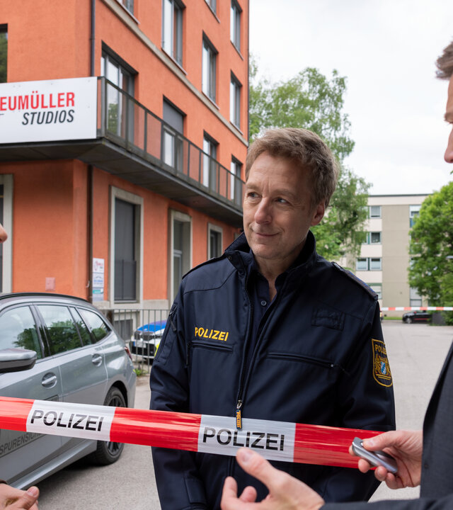 "Die Rosenheim-Cops - Die Autogrammjagd": Kommissar Kaya (Baran Hêvî) und Hauptkommissar Hansen (Igor Jeftić) stehen nebeneinander vor rotweißem Flatterband mit der Aufschrift "Polizei" und sprechen mit Polizeihauptmeister Mohr, der hinter dem Absperrband steht und Kaya ansieht. Im Hintergrund steht ein rötliches Gebäude, an dem ein Schild mit der Aufschrift "Neumüller Studios" angebracht ist.