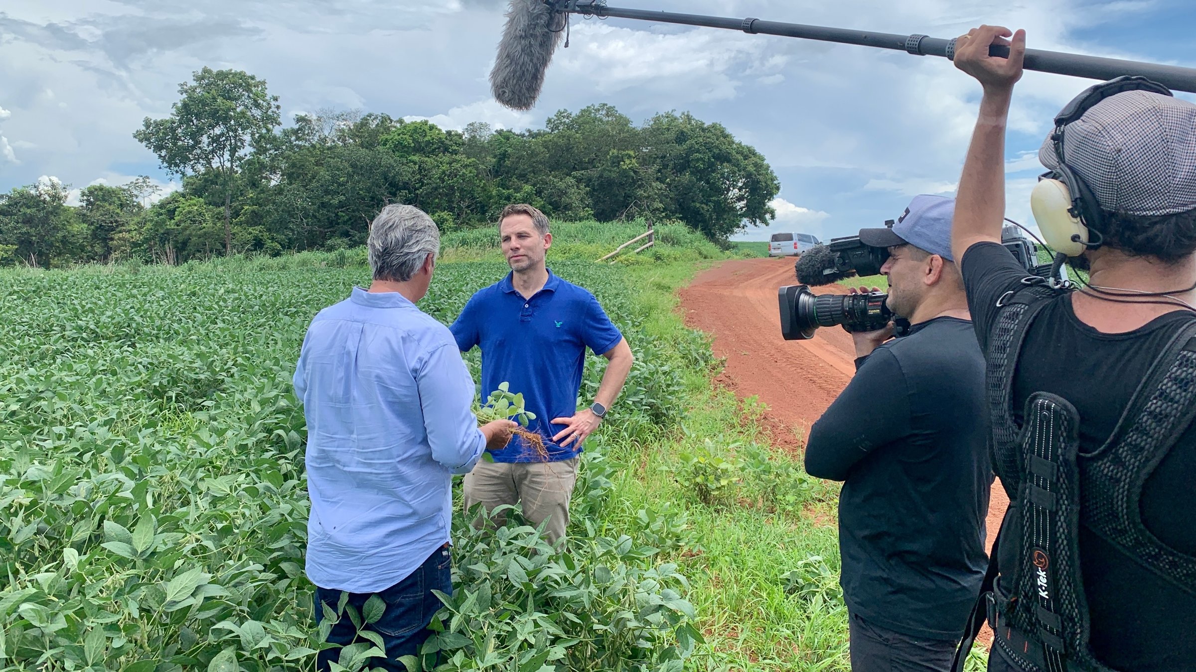 "auslandsjournal - die doku: Zeitenwende global - Russlands Angriff auf die Freiheit": Der Südamerika-Korrespondent Christoph Röckerath und der Agrarunternehmer Osvaldo Pasqualotto stehen in einem Feld und sprechen miteinander. Osvaldo Pasqualotto sieht man nur von hinten. Rechts im Bild sieht man einen Kameramann und einen Tontechniker bei der Arbeit.