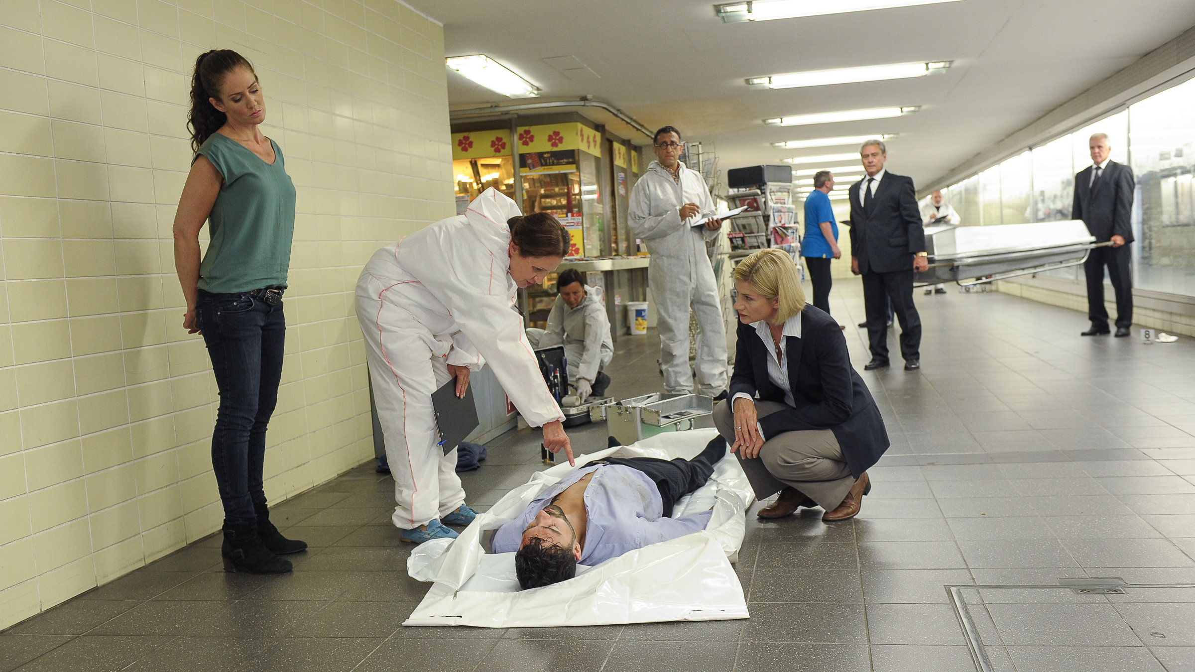 "SOKO Stuttgart - Der Held von Stuttgart": Prof. Lisa Wolter (Eva Maria Bayerwaltes) zeigt Martina Seiffert (Astrid M. Fünderich) etwas auf der Brust des Opfers Daniel Pflüger (Komparse). Selma Kirsch (Yve Burbach) steht daneben und hört zu. Im Hintergrund steht Jan Arnaud (Mike Zaka Sommerfeldt), der die Situation beobachtet sowie Spurensicherer (Komparsen), die die Umgebung absuchen. Die Bestatter (Komparsen) tragen den Sarg zur Leiche.