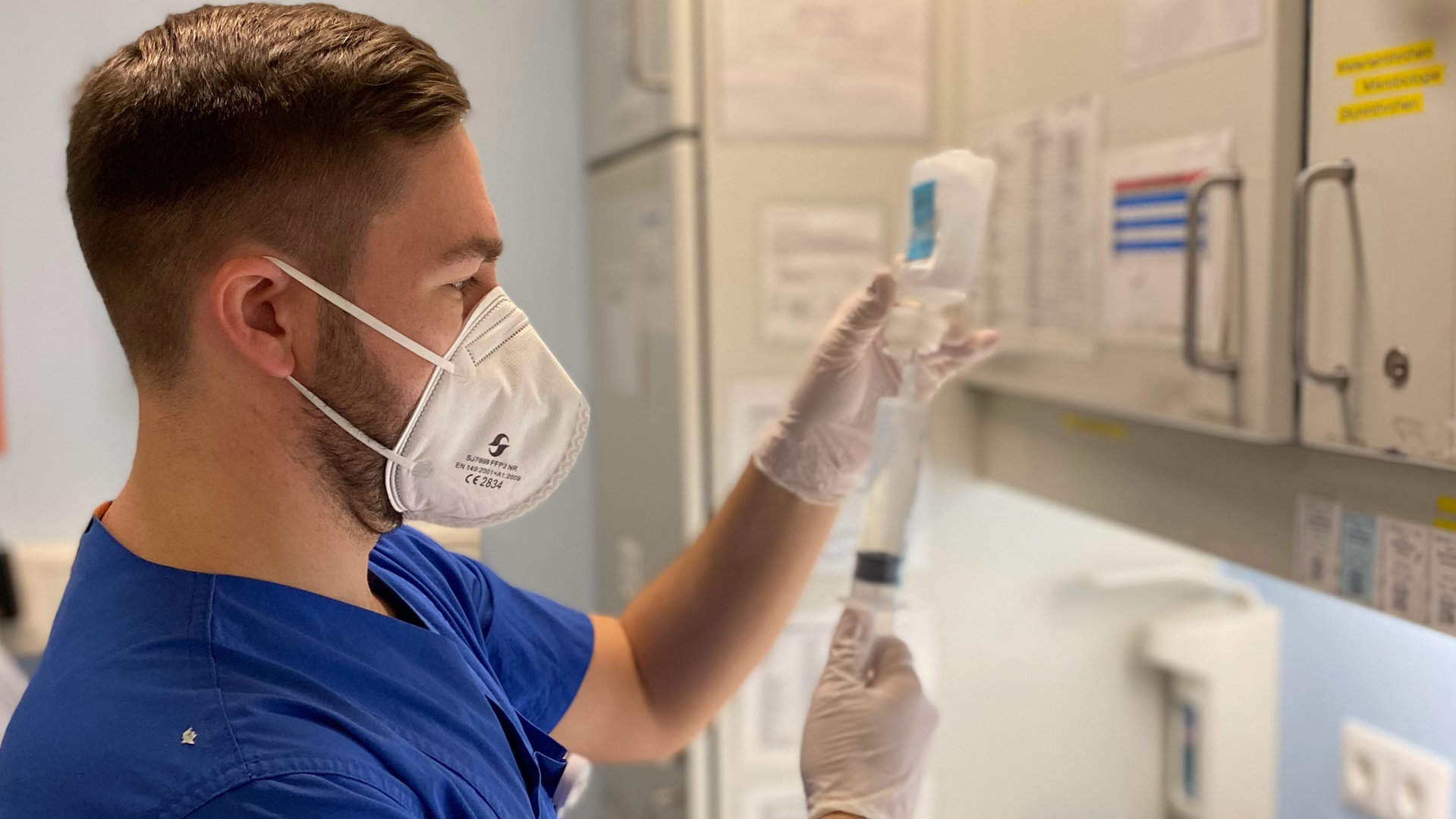 "Leben ist mehr! Intensivpflege – ein täglicher Kampf ums Leben": Dominik Stark, ein junger Mann, zieht ein Medikament in eine Spritze auf. Er steht in der Notfallstation einer Uni-Klinik, trägt eine weiße FFP2 Maske und ein blaues Oberteil.
