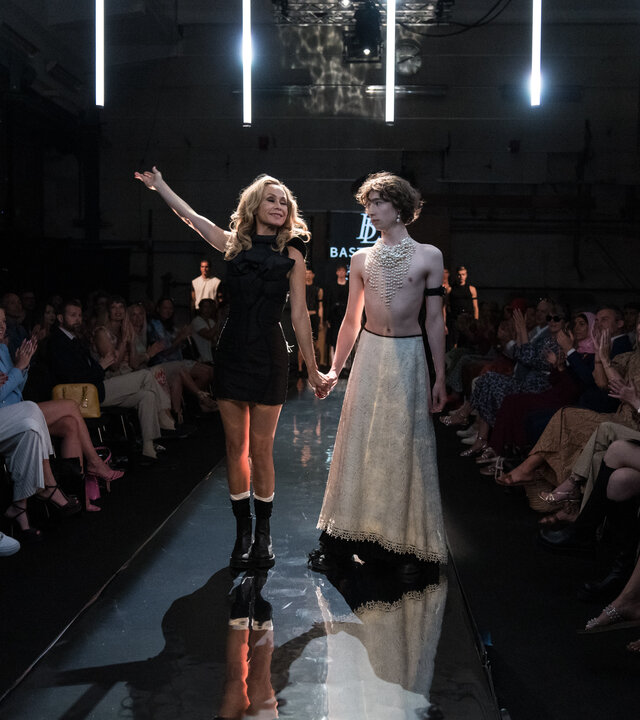 "Dan Sommerdahl – Tödliche Idylle: Fashion Week": Bastienne (Kristina Korsholm) steht mit Model Jonathan (Oskar Ruskjær), der sie an der Hand hält auf dem Laufsteg. Sie hebt den rechten Arm und blickt triumphierend nach oben, während Jonathan sie zurückhaltend ansieht. Das Publikum (Komparsen) sitzt zu beiden Seiten des Laufstegs und applaudiert.