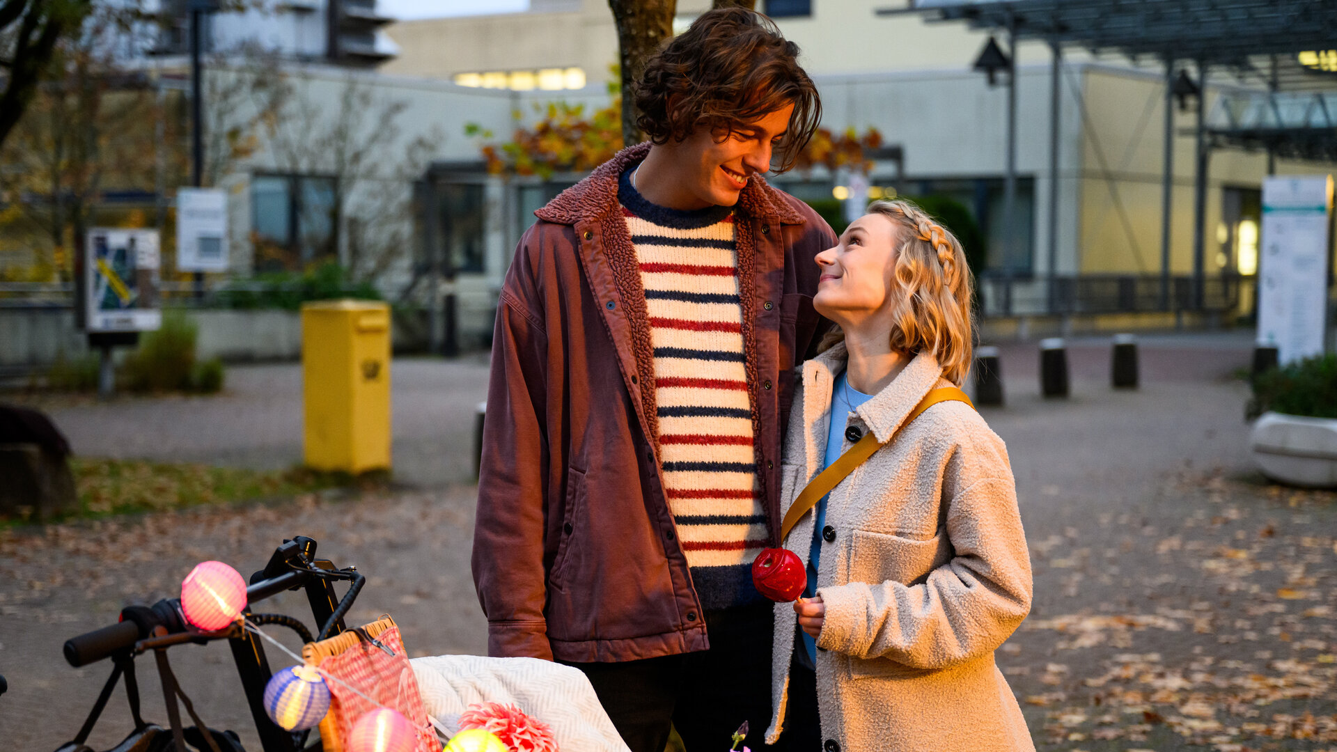 "Bettys Diagnose - Spielereien": Auf einem kleinen Platz in der Stadt: Simon (Ivan Barišić) hält Mia (Antonia Döring) im Arm und schaut sie verliebt an. Im Vordergrund steht ein Lastenfahrrad mit einer Lichtergirlande, am Lenker hängt ein Fahrradhelm. Im Hintergrund steht ein großer, gelber Briefkasten.