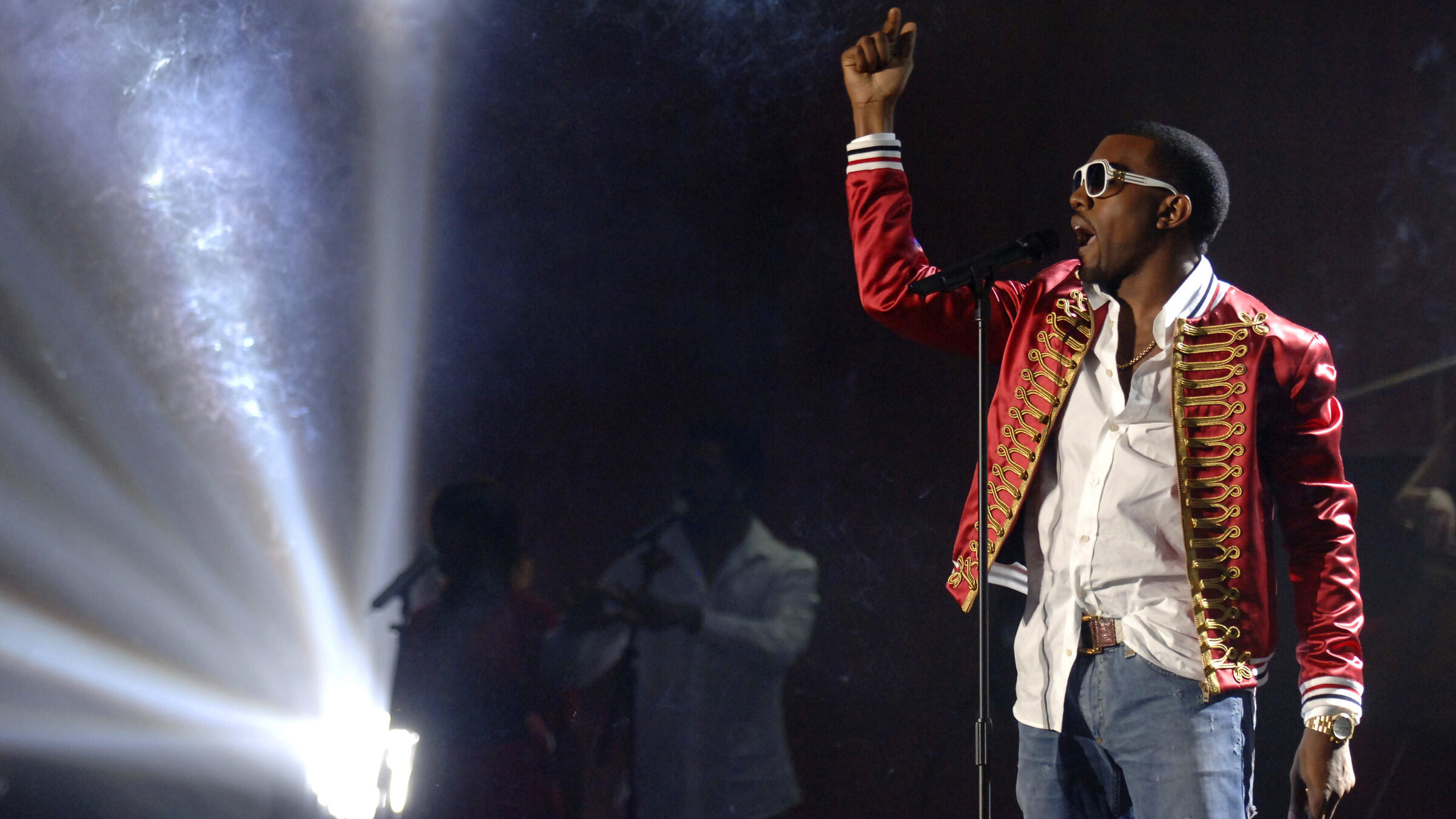 "The True Story of Kanye West": Bühne mit grellem Scheinwerferlicht links. Rechts ein Mann in Jeans, weißem Hemd und rotem Zirkus-Sakko, trägt Sonnenbrille