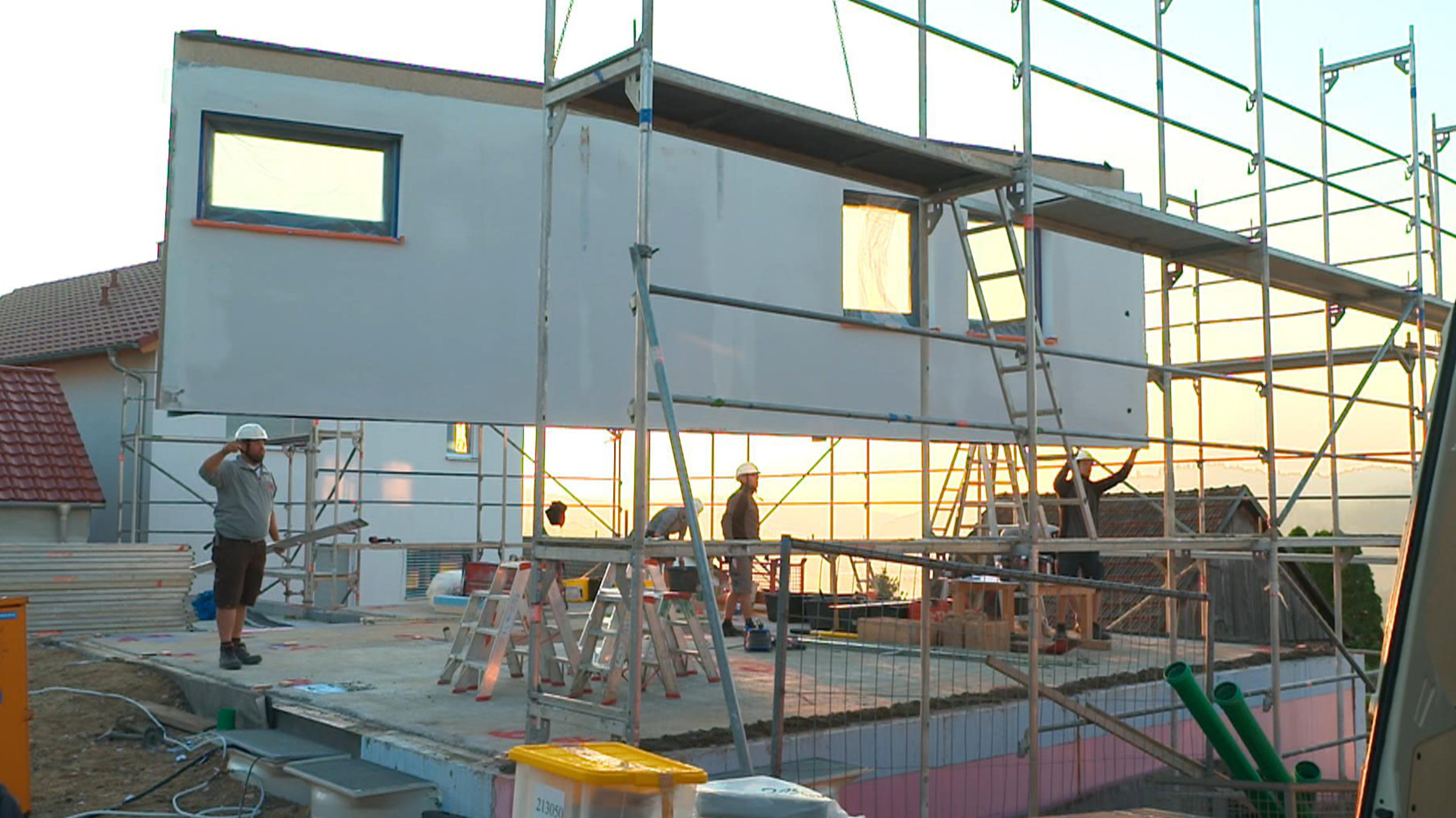 "ZDF.reportage: Albtraum Hausbau? - Steine, Stress und Schulden": Bauarbeiter setzen auf einer Bodenplatte ein Wandteil in Holztafelbauweise an. Das Wandteil schwebt in der Luft gehalten von einem Kran. Die Sonne geht im Hintergrund auf. Im Vordergrund steht ein Gerüst.