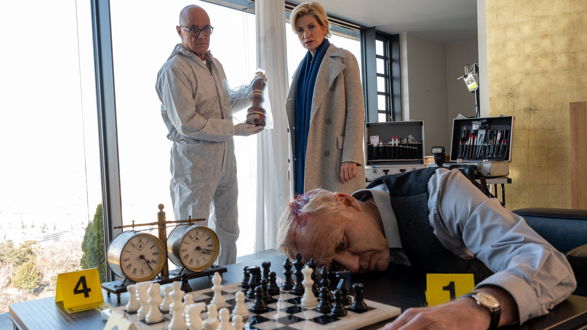 "SOKO Stuttgart - Schachmatt": Kriminalhauptkommissarin Martina Seiffert (Astrid M. Fünderich) und ein Spurensicherer (Komparse) stehen am Tatort. Sie schauen auf die Leiche: ein Mann, der mit dem Oberkörper auf einem Tisch neben einem Schachbrett liegt. An seinem Kopf ist eine klaffende Platzwunde zu sehen. Der Spurensicherer hält einen Schachpokal in einer Asservatentüte in den Händen.