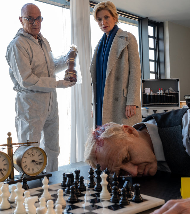 "SOKO Stuttgart - Schachmatt": Kriminalhauptkommissarin Martina Seiffert (Astrid M. Fünderich) und ein Spurensicherer (Komparse) stehen am Tatort. Sie schauen auf die Leiche: ein Mann, der mit dem Oberkörper auf einem Tisch neben einem Schachbrett liegt. An seinem Kopf ist eine klaffende Platzwunde zu sehen. Der Spurensicherer hält einen Schachpokal in einer Asservatentüte in den Händen.