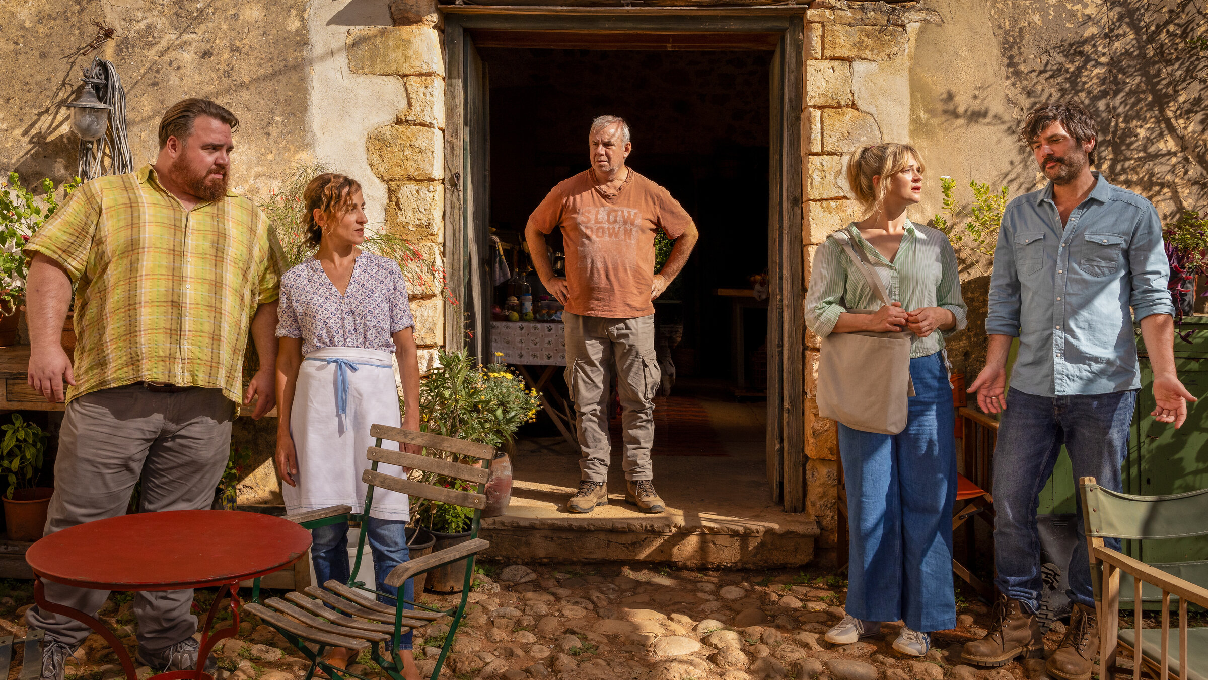"Endlich Witwer - Griechische Odyssee": Verwirrt steht Georg Weiser (Joachim Król) nach einem schief gelaufenen Familientreffen mit seinen beiden Kindern in einem großen Türrahmen eines Steinhauses auf Kreta. Rechts im Bild steht seine Tochter Susanne (Caroline Hanke) neben ihrem Partner Alex (Jasin Challah). Links vor der Hauswand steht sein Sohn Gerd (Tristan Seith) neben Alex‘ Frau Helena (Artemis Chalkidou).