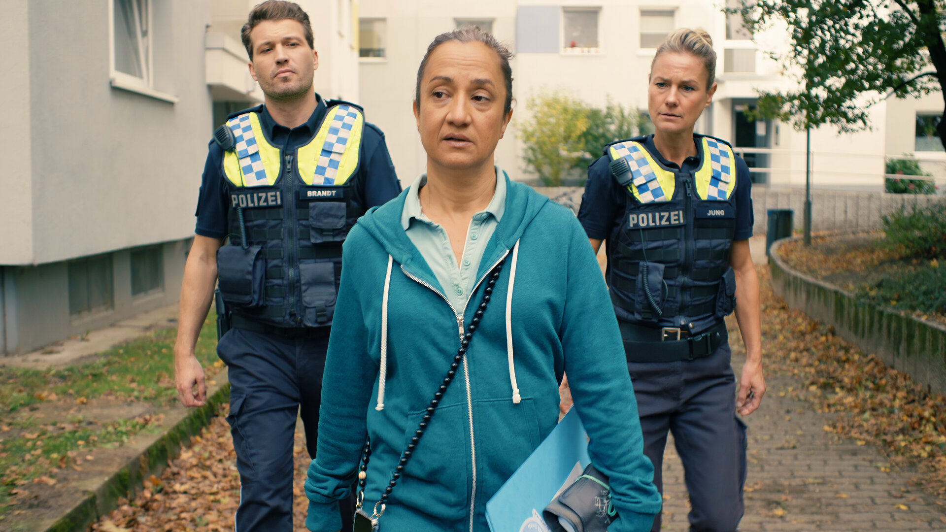 "Notruf Hafenkante - Überrollt": Der Polizist Nick Brandt (Raúl Richter) und seine Kollegin Franzi Jung (Rhea Harder-Vennewald) laufen nebeneinander hinter Susann Peters (Meriam Abbas) einen Weg vor einer Wohnanlage entlang. Beide blicken fragend zu Susann.