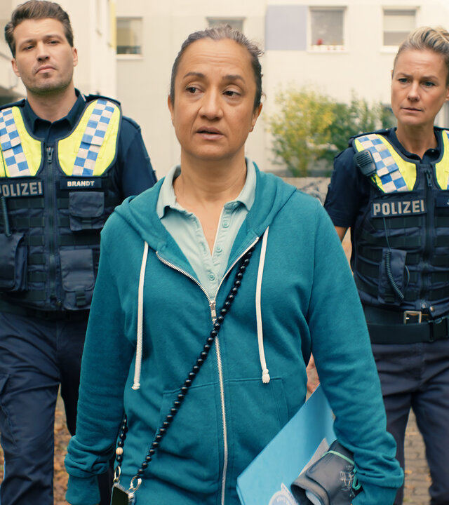 "Notruf Hafenkante - Überrollt": Der Polizist Nick Brandt (Raúl Richter) und seine Kollegin Franzi Jung (Rhea Harder-Vennewald) laufen nebeneinander hinter Susann Peters (Meriam Abbas) einen Weg vor einer Wohnanlage entlang. Beide blicken fragend zu Susann.