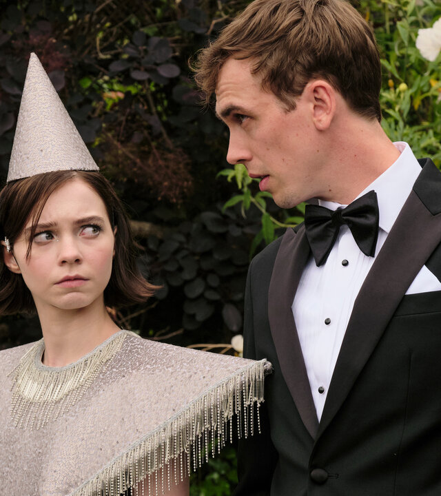 "A Good Girl's Guide to Murder - Schulprojekt: Mord": Pip (Emma Myers) befindet sich auf einer Party in einem unangenehmen Gespräch mit Dan Da Silva (Jackson Bews). Sie trägt einen gräulichen Fransenponcho und einen spitzen Partyhut. Er trägt einen schwarzen Anzug mit Fliege. Während sie von links argwöhnisch zu ihm hochschaut, redet er  angeregt auf sie ein.