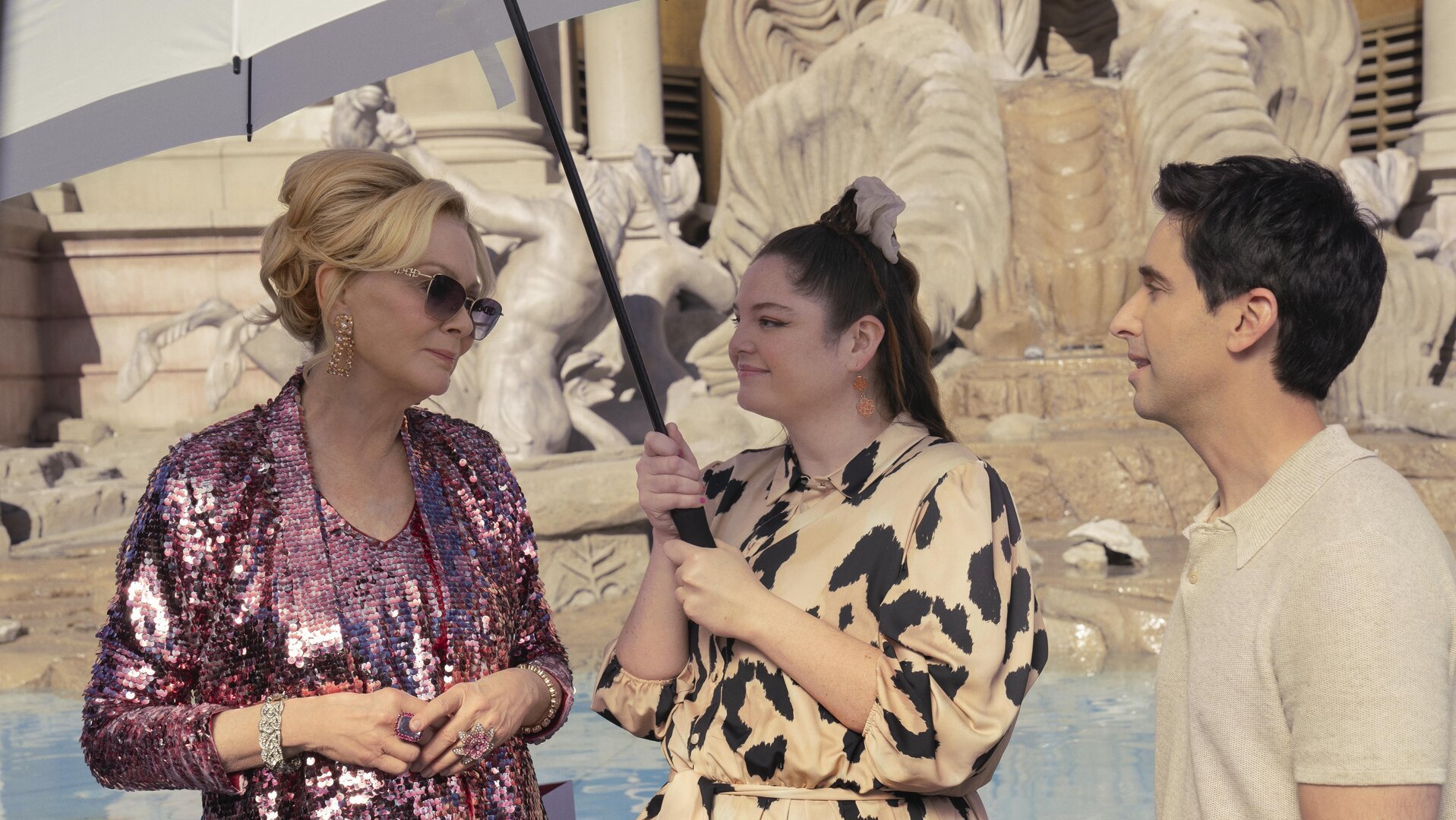 "Hacks - Besser spät": Deborah (Jean Smart, l.) in einem pinken Pailettenlook steht neben Kayla (Meghan Stalter), die schützend einen Regenschirm als Sonnenschutz über sie hält, und Jimmy (Paul W. Downs). Im Hintergrund ist der Nachbau des Trevi-Brunnens zu erkennen.