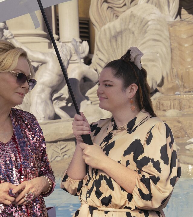 "Hacks - Besser spät": Deborah (Jean Smart, l.) in einem pinken Pailettenlook steht neben Kayla (Meghan Stalter), die schützend einen Regenschirm als Sonnenschutz über sie hält, und Jimmy (Paul W. Downs). Im Hintergrund ist der Nachbau des Trevi-Brunnens zu erkennen.