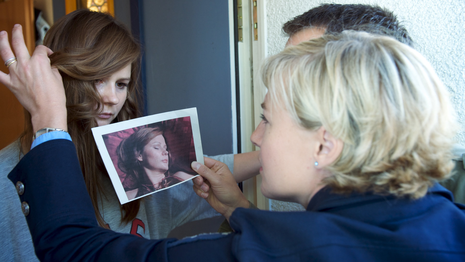 "SOKO Wismar Böses Erwachen": Janine Franzen (Annett Heilfort) steht in einer offenen Wohnungstür. Vor ihr vergleichen Leena Virtanen (Li Hagman) und Sven Herzog (Michael Härle) die Frau auf einem Foto mit ihr. Leena hält eine Strähne aus Janines Gesicht.