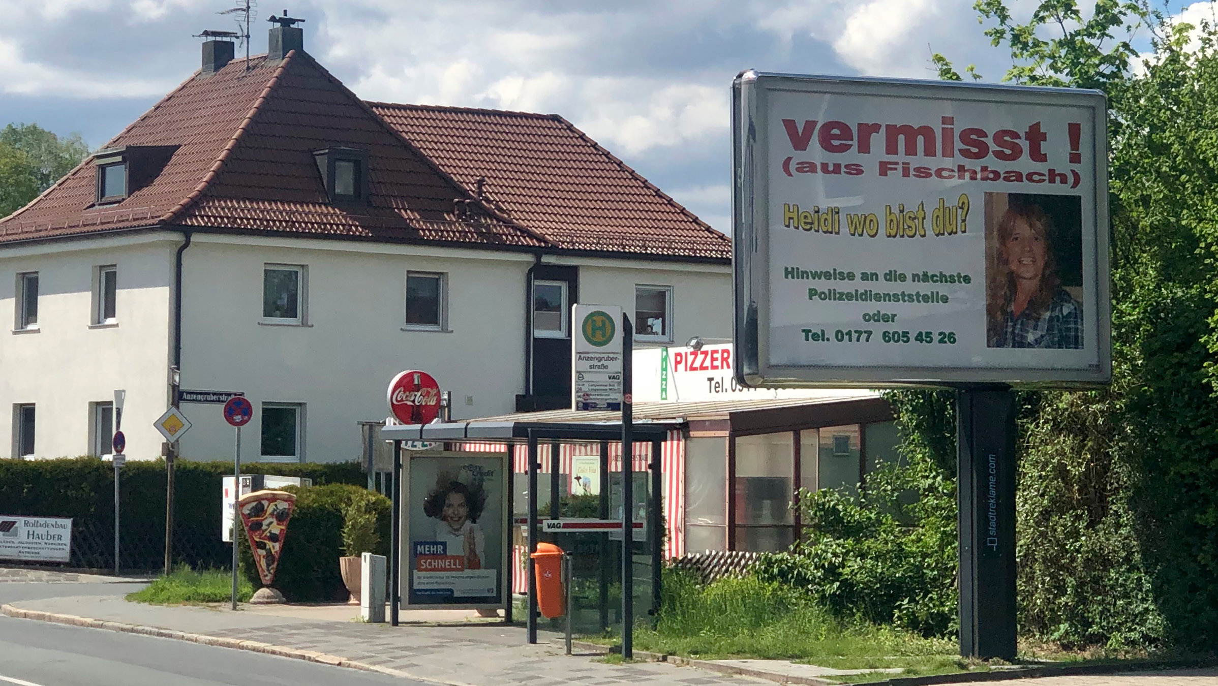 "37°: Vermisst - Wenn Menschen spurlos verschwinden": Ein riesiges Plakat neben einer Bushaltestelle. Auf dem Plakat ist ein Foto zu sehen mit einer blonden Frau. Darauf der Titel 'Heidi wo bist du'? Im Hintergrund eine unbefahrene Straße und links im Bild ein Haus.