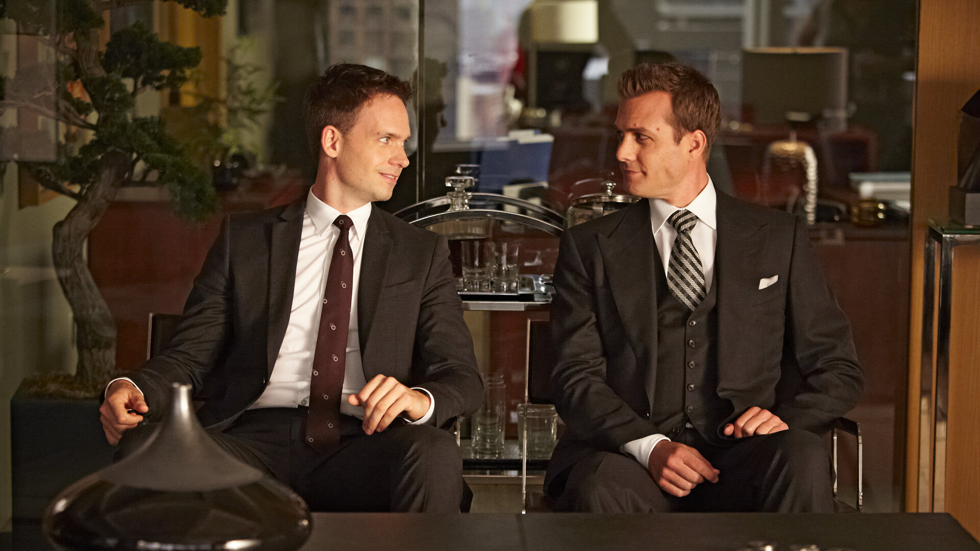 "Suits - Offene Rechnung:" Harvey (Gabriel Macht) und Mike (Patrick J. Adams) sitzen nebeneinander in einem modern eingerichteten Büro. Die beiden blicken sich lächelnd an. Im Hintergrund befindet sich ein Bonsai-Baum, im Vordergrund steht eine Karaffe auf einer Tischplatte.