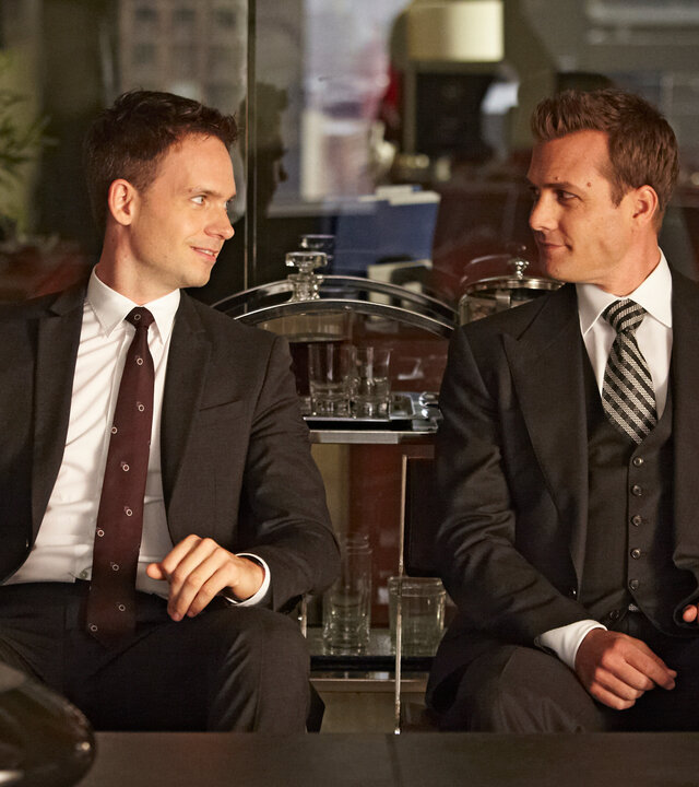 "Suits - Offene Rechnung:" Harvey (Gabriel Macht) und Mike (Patrick J. Adams) sitzen nebeneinander in einem modern eingerichteten Büro. Die beiden blicken sich lächelnd an. Im Hintergrund befindet sich ein Bonsai-Baum, im Vordergrund steht eine Karaffe auf einer Tischplatte.