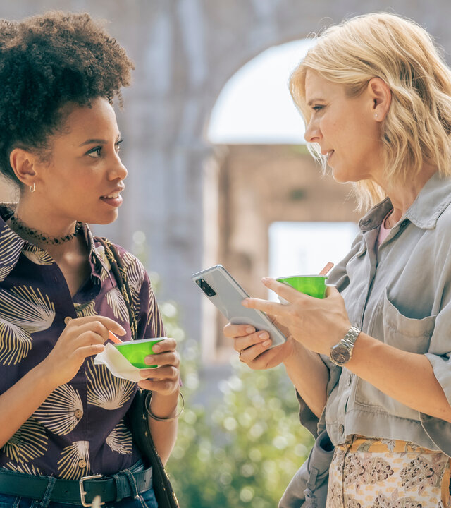 "Signora Volpe - Verkannte Helden": Raffaella (Marianne Leoni) und Sylvia (Emilia Fox) stehen sich vor dem Kolosseums in Rom gegenüber. Beide halten Eisbecher in der Hand, Sylvia zudem noch ihr Mobiltelefon.