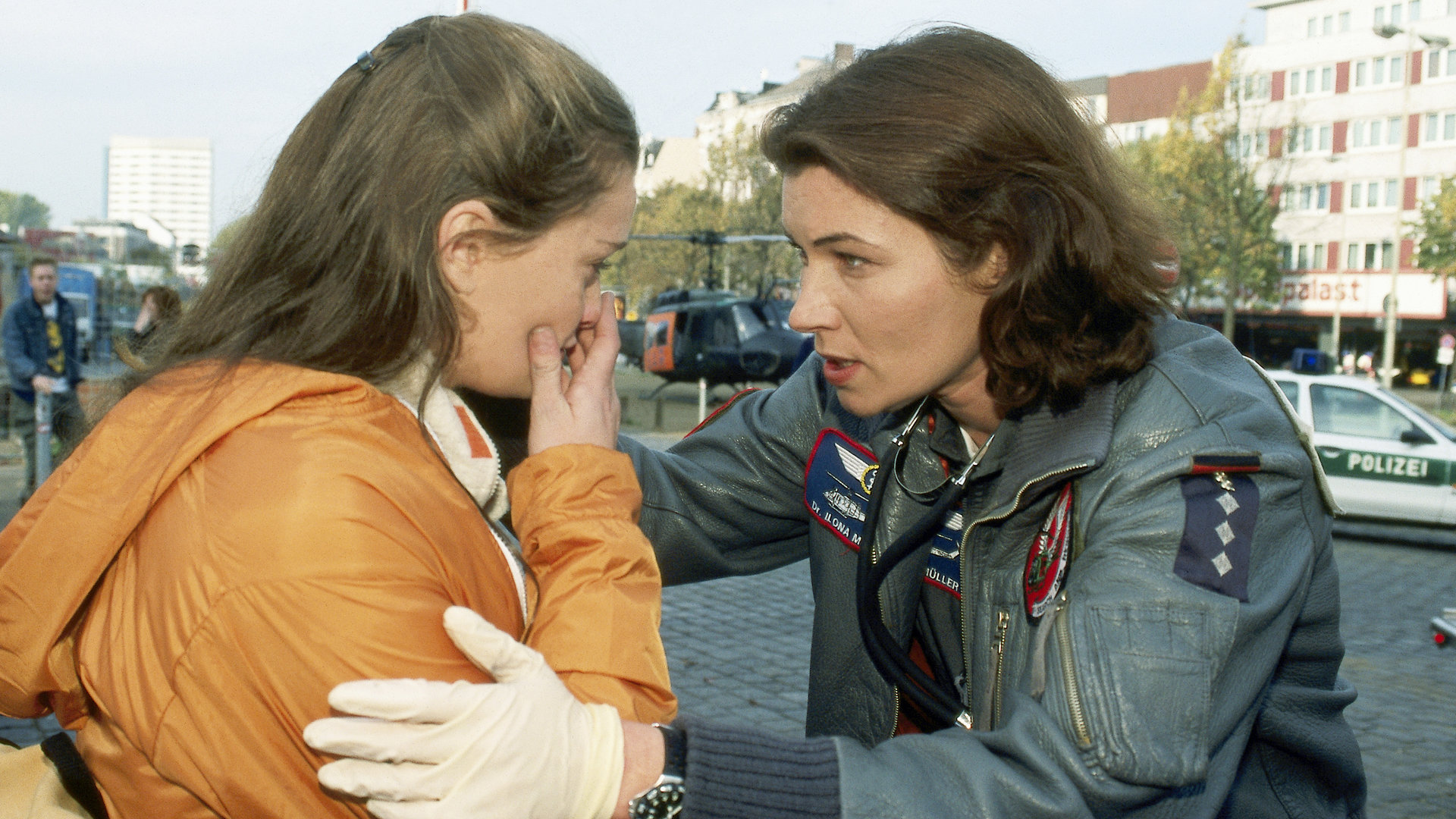 "Die Rettungsflieger - Explosiv": Straßenszene. Im Hintergrund stehen der Rettungshubschrauber und Polizeifahrzeuge. Andrea (Natalie Spinel) und Ilona (Julia Heinemann) stehen sich gegenüber. Ilona neigt sich Andrea zu und spricht beruhigend auf sie ein. Andrea hält sich entsetzt die Hand vor den Mund.